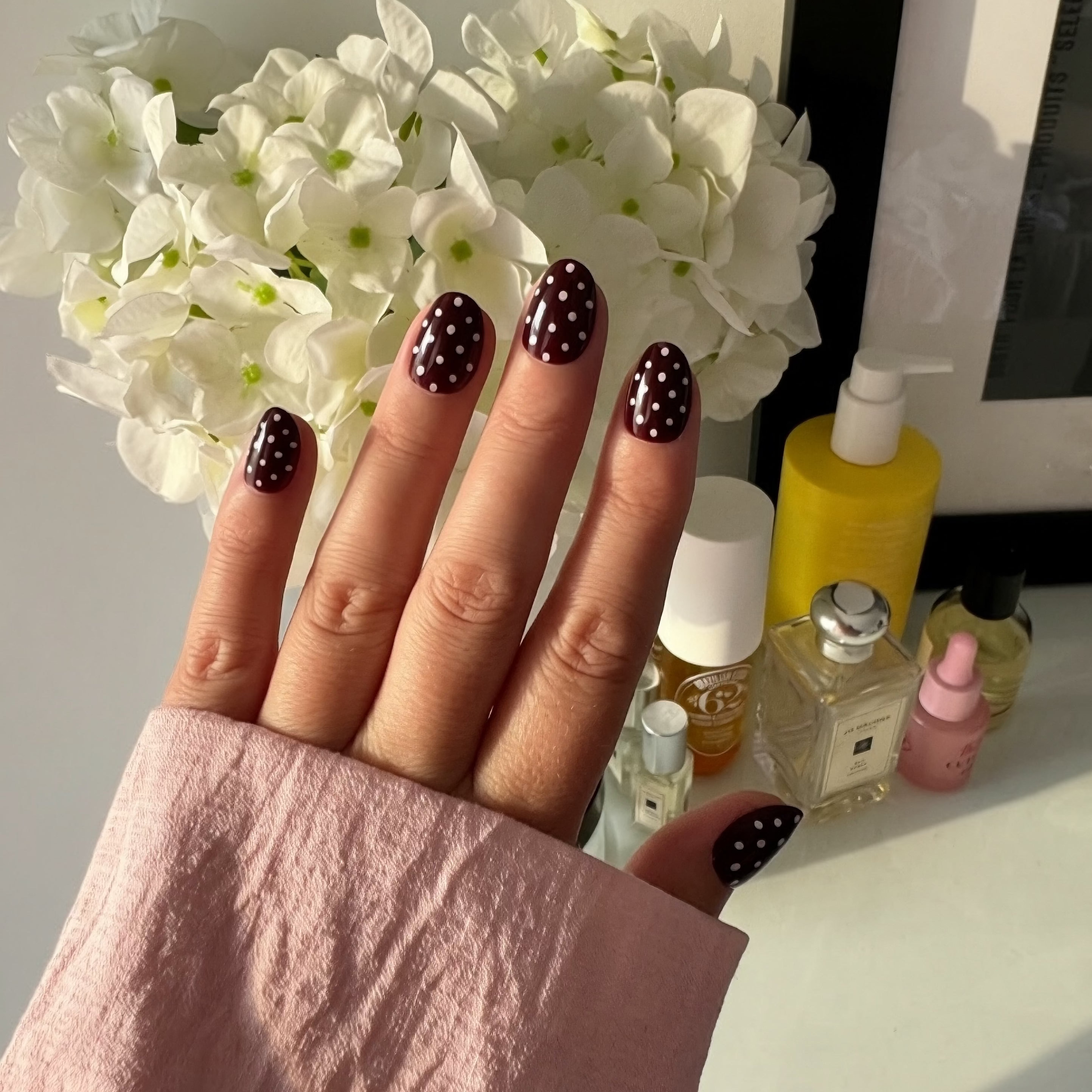 Hand with dark press polka dot Instant Mani Co. press on nails with skin care in background