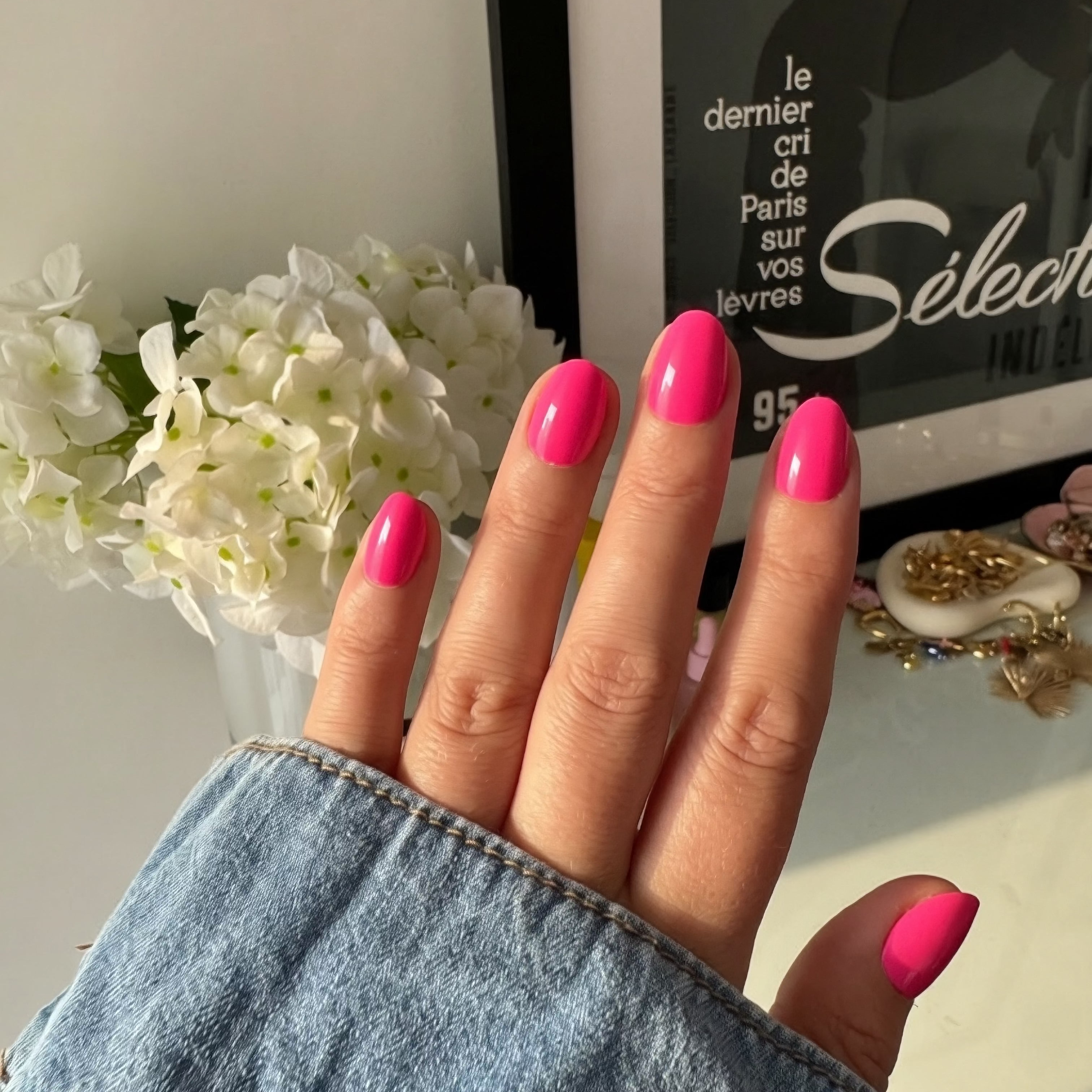 Hand with Instant Mani Co. hot pink Barbie nails holding a denim jacket, with a blurred background of flowers and a product display.