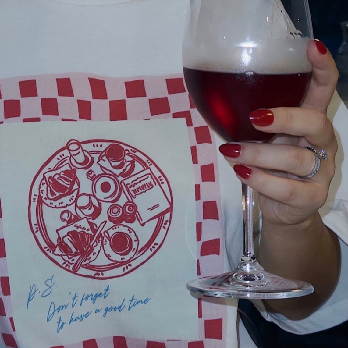 Person wearing a white t-shirt with a red and green graphic design, holding a glass of red wine. And wearing Instant Mani Co. Ruby Red press on nails