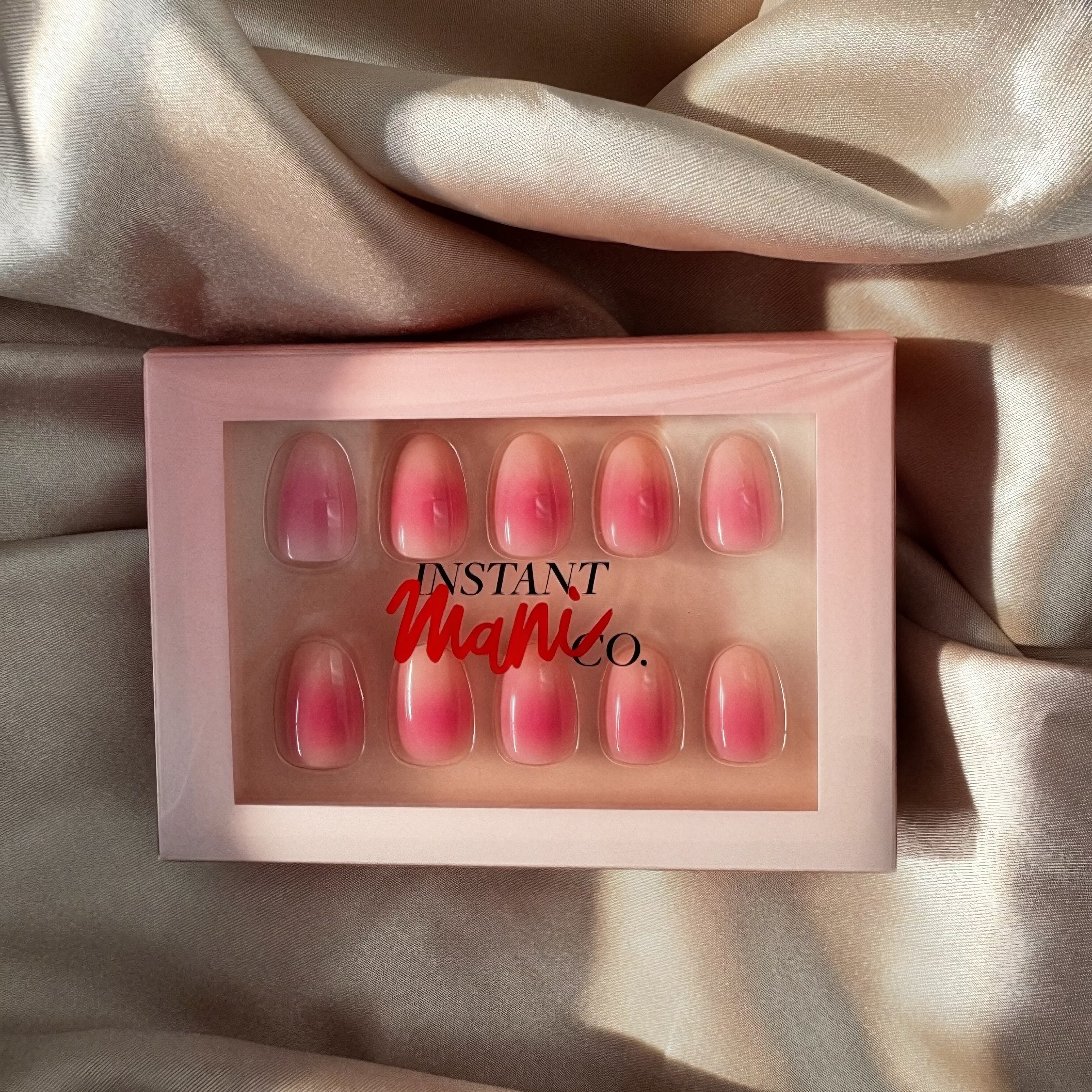 A set of 10 pink press-on nails with a gradient design, displayed in a light pink box with the brand name 'Instant Mani Co.' printed on the front, set against a draped champagne-colored satin fabric.