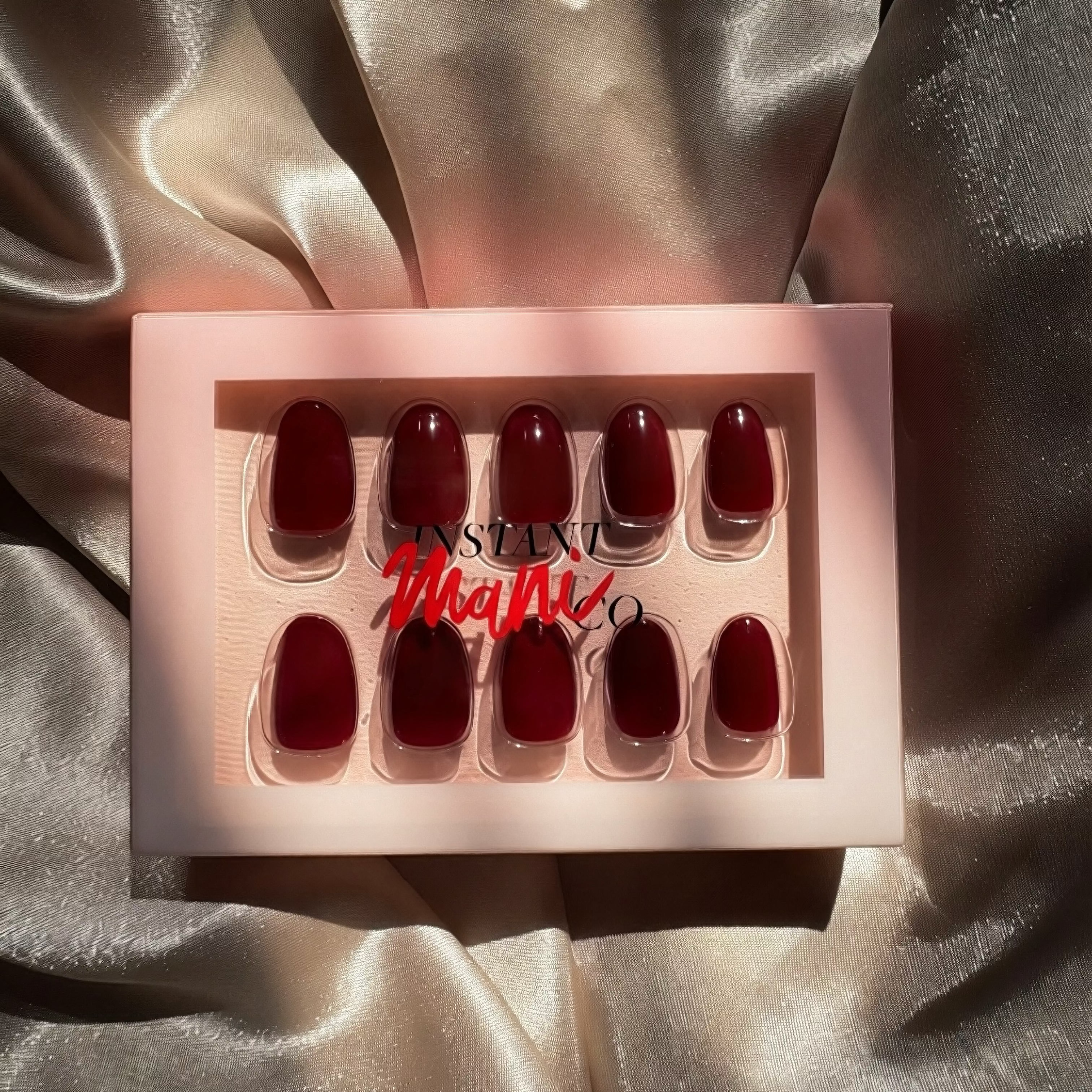 A set of Instant Mani Co. press-on nails in a deep burgundy red shade, presented in a light pink box with a clear window. The box has a red 'INSTANT Mani Co' logo overlay.