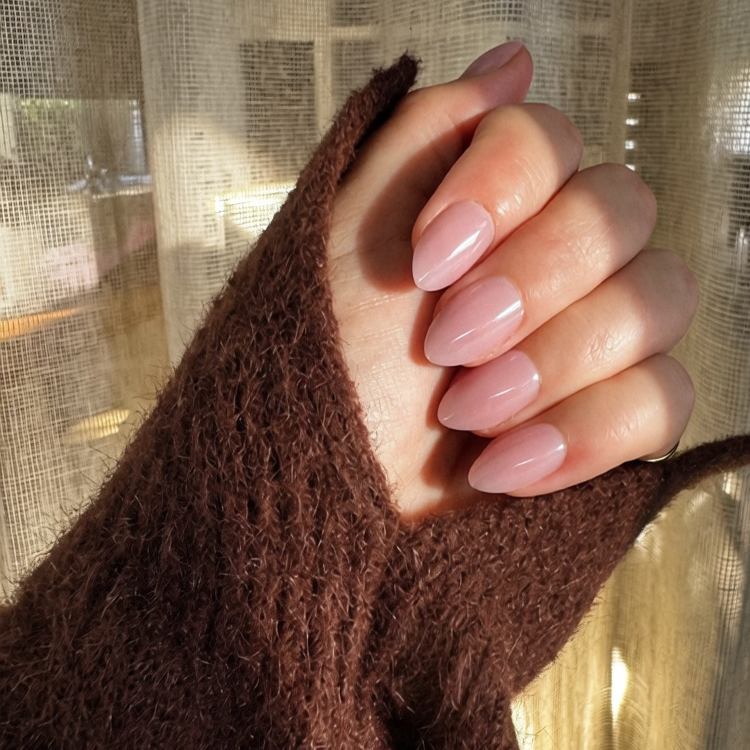 A close-up of a hand with Instant mani Co. almond-shaped press-on nails in a glossy dusty pink shade called Halo, wearing a brown knit sweater sleeve against a background of sheer beige curtains with warm sunlight.