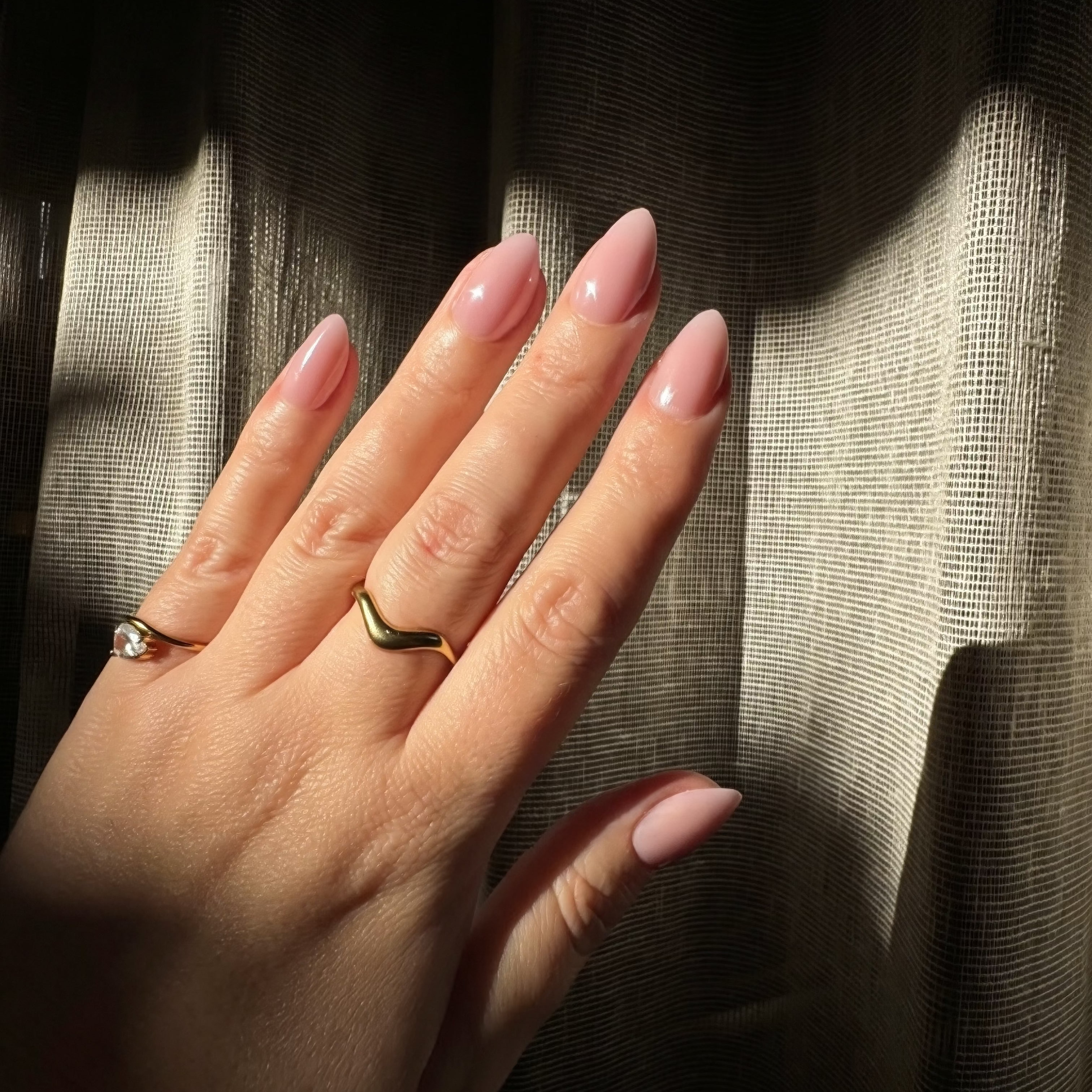 A close-up of a hand with Instant mani Co. almond-shaped press-on nails in a glossy dusty pink shade called Halo, wearing a brown knit sweater sleeve against a background of sheer beige curtains with warm sunlight.