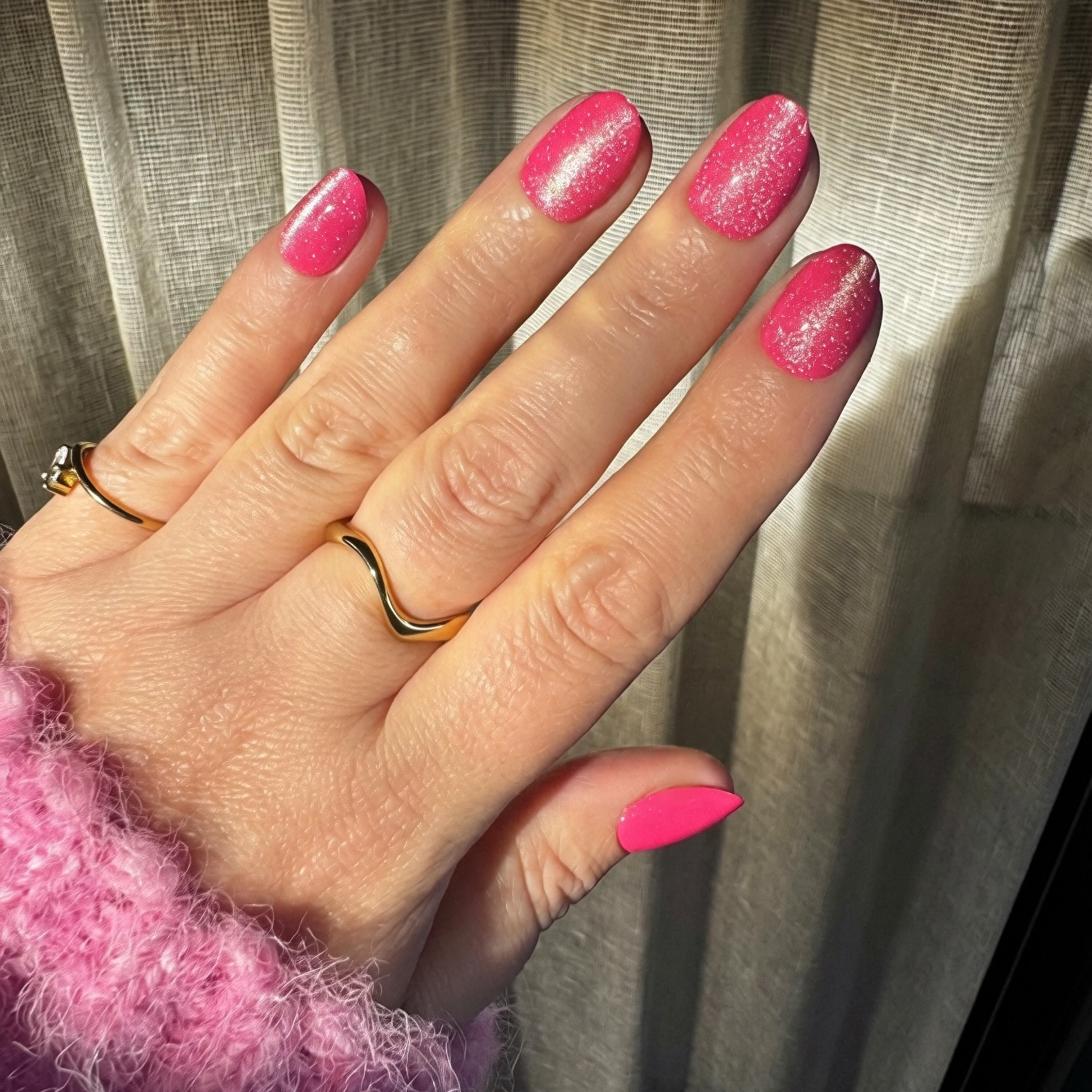 A close-up of a hand with bright velvet Instant Mani Co. press on nails, wearing gold rings and a fuzzy pink sweater, with sheer curtains in the background.