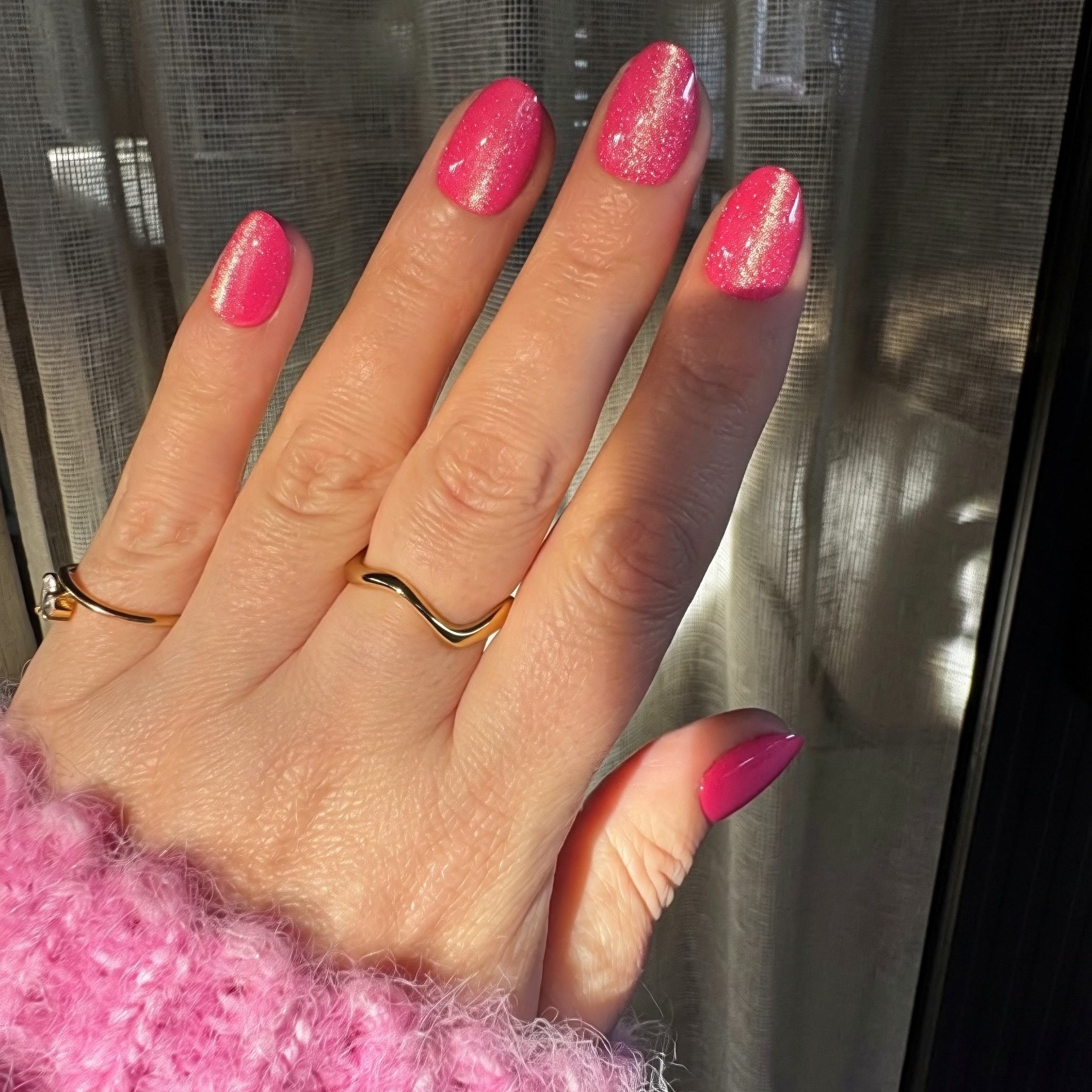 A close-up of a hand with bright velvet Instant Mani Co. press on nails, wearing gold rings and a fuzzy pink sweater, with sheer curtains in the background.