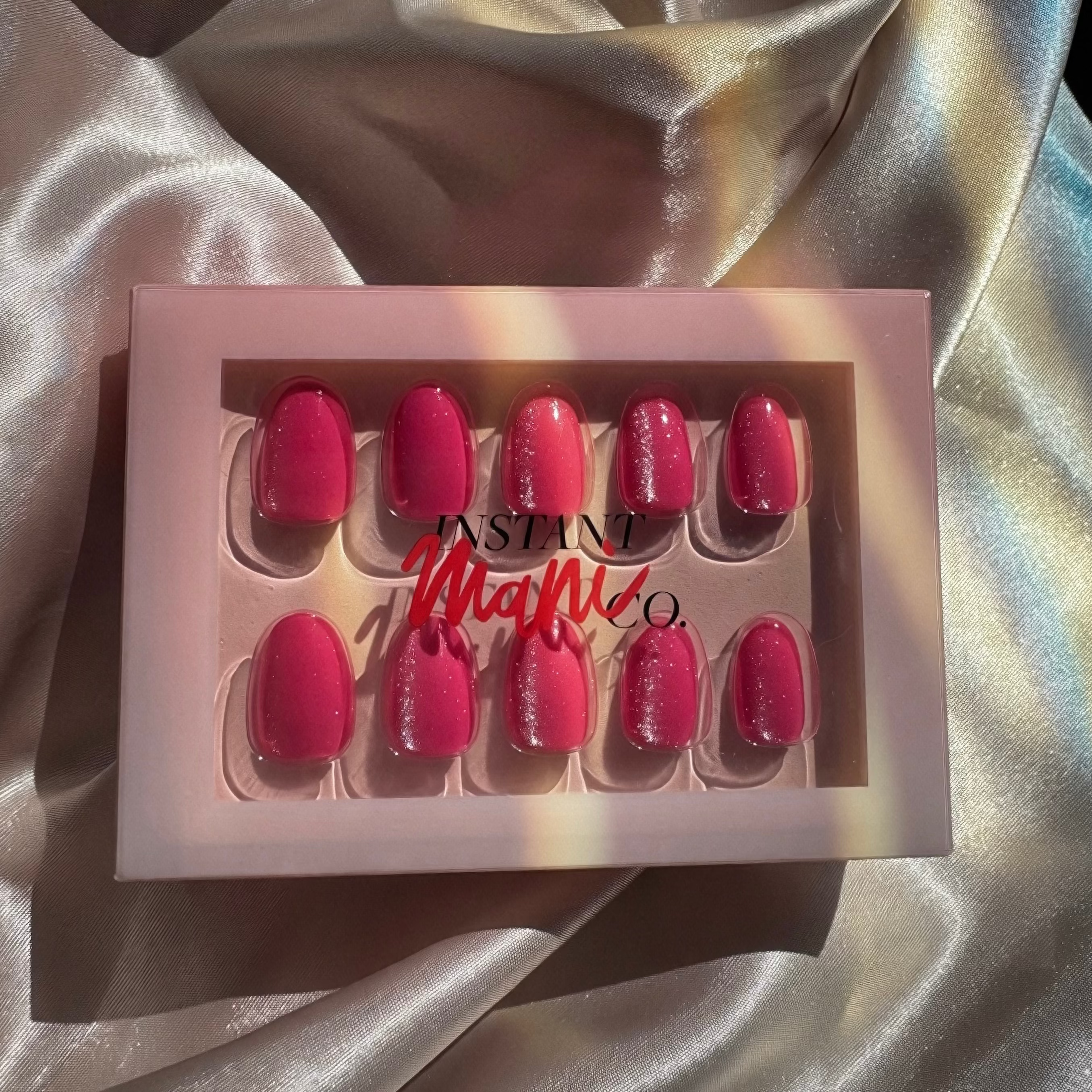 A set of Instant Mani Co Velvet pink press-on nails in a light pink box with the brand name 'Instant Mani Co.' printed on the front, styled on a shiny champagne satin fabric background.