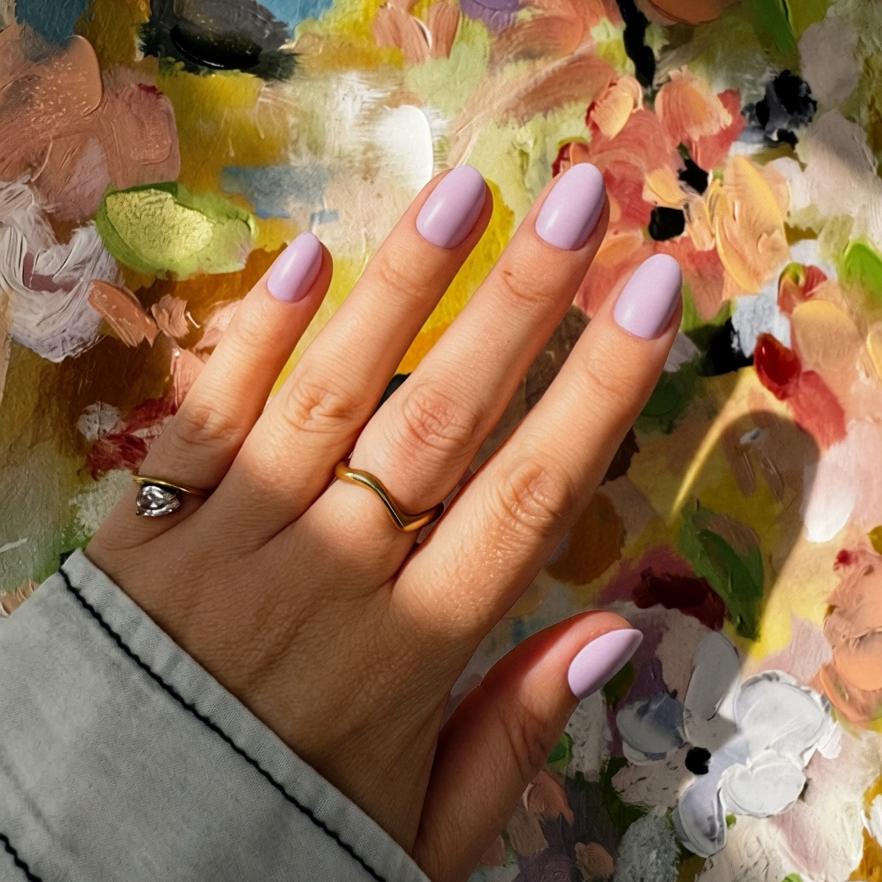 A close-up of a hand with Instant Mani Co. Lilac Haze press on nails resting on a colorful, textured oil painting palette. The hand is wearing a gold ring and a thin gold band.