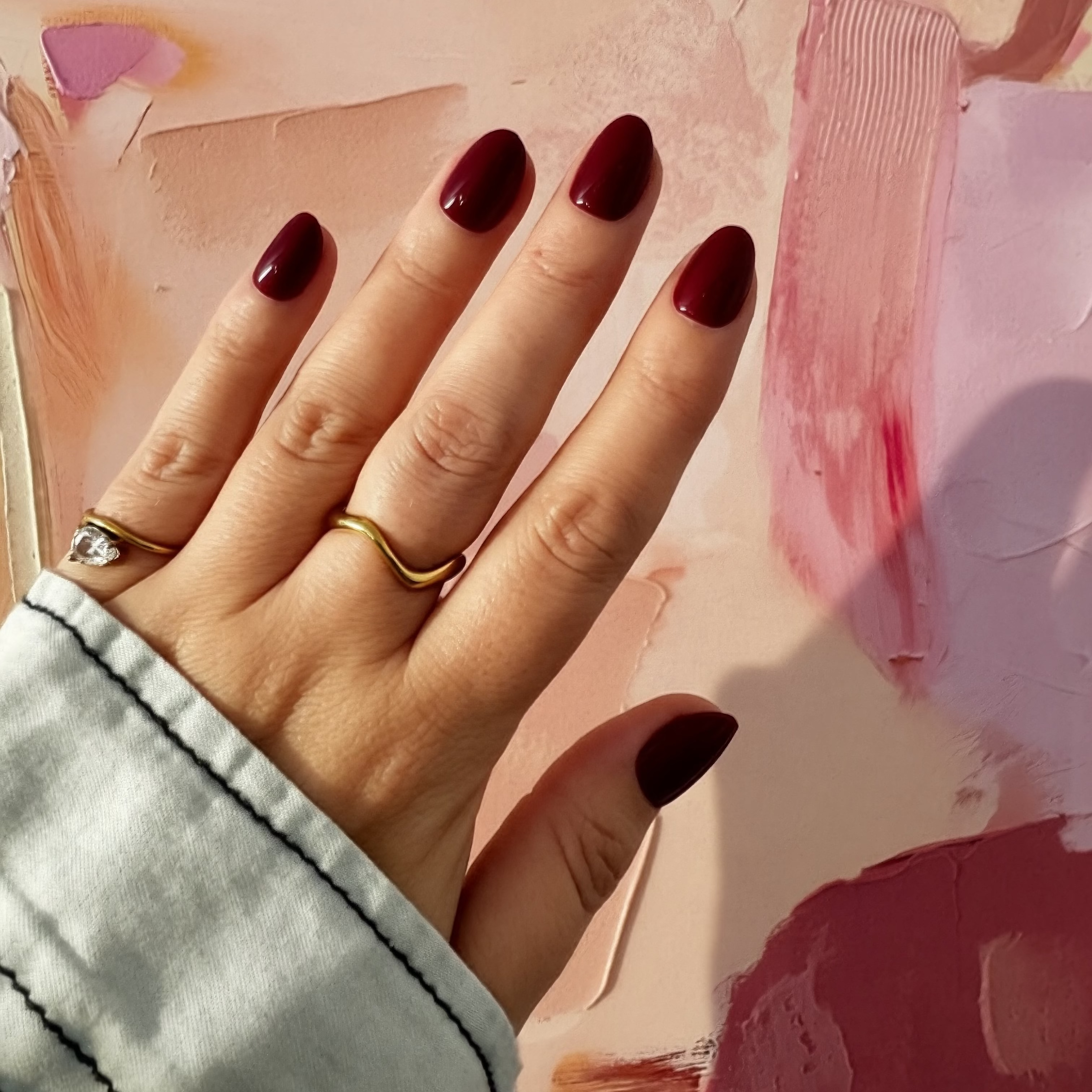 A close-up of a hand with glossy Instant Mani Co. burgundy Merlot press on nails, wearing two gold rings and a light denim sleeve, posed against an abstract pink and peach painted background.