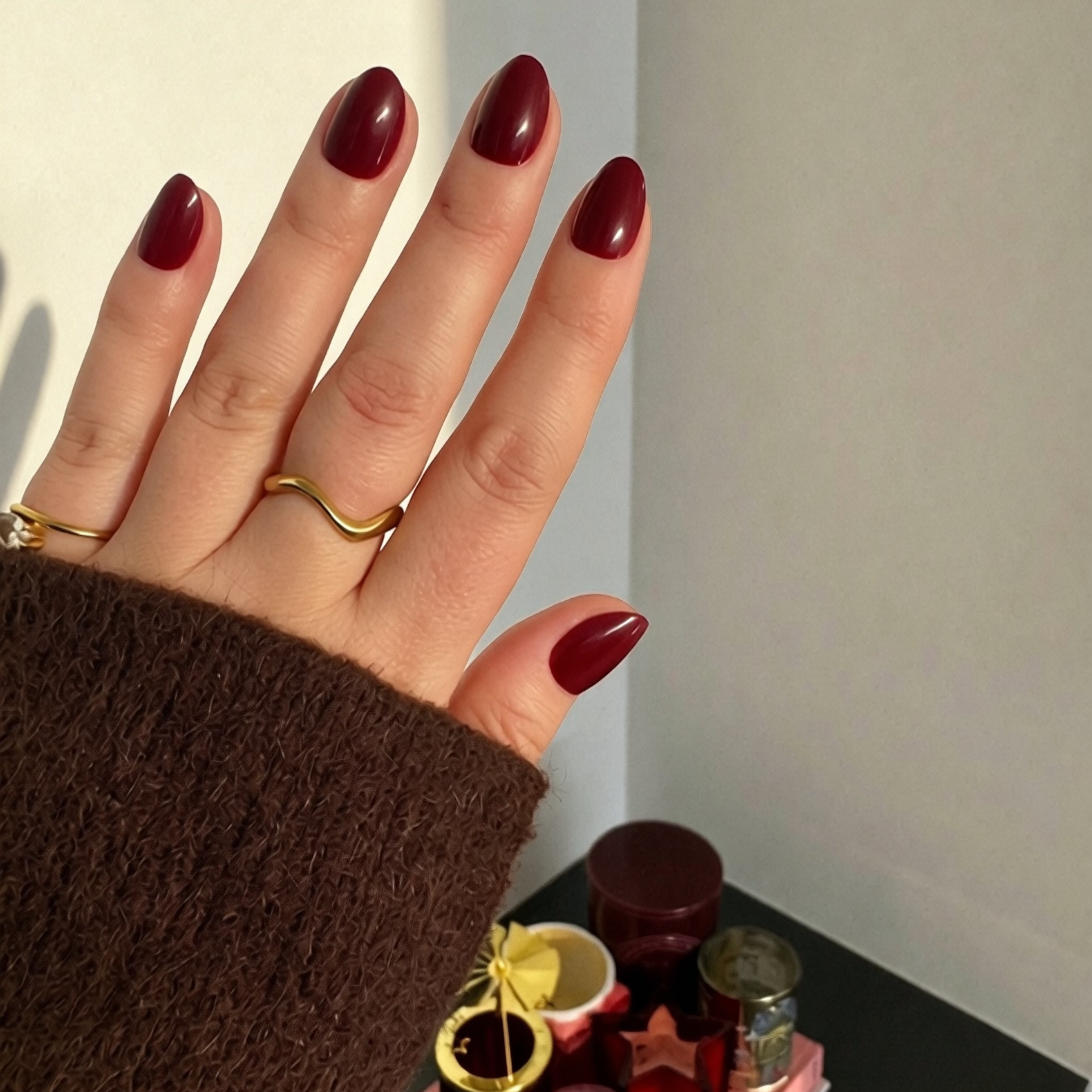 A close-up of a hand with glossy Instant Mani Co. burgundy Merlot press on nails, wearing two gold rings and a brown sleeve, posed against a candle display