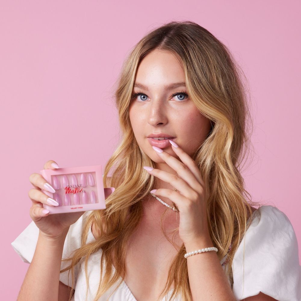Girl wearing Instant Mani Co Velvet Pink Press on nails, holding Instant Mani Co. box, standing in front of pink background wearing white shirt