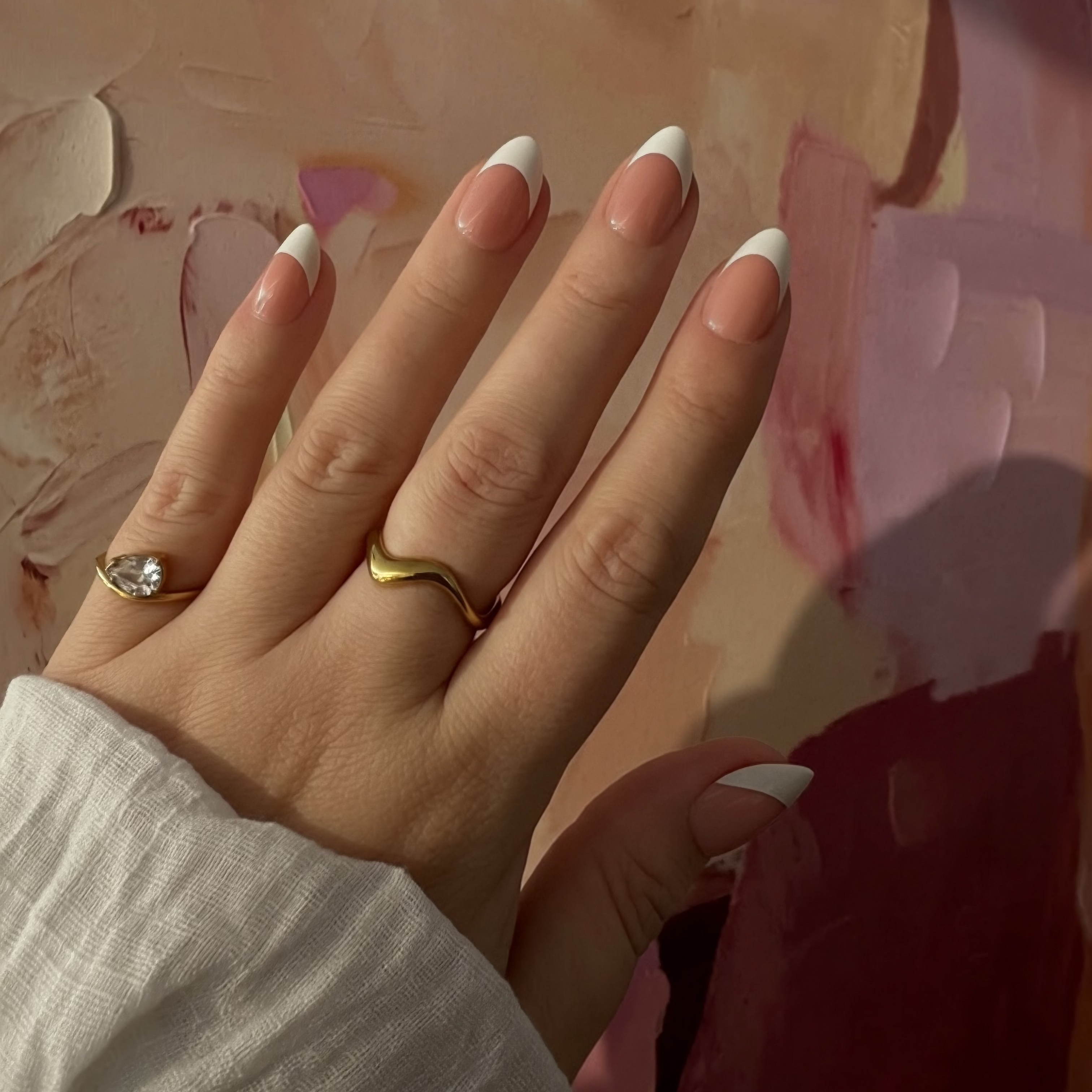 A close-up of a hand with French Instant Mani Co. Press on nails wearing two rings: a gold band with a small clear gemstone and a gold wave ring, set against a pink textured background