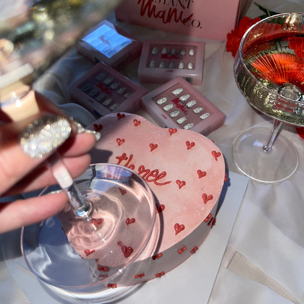 Girl wearing sparkly nails. holding champaign, in front of pink heart cake, with Lover Instant Mani Co press on nails in background. 