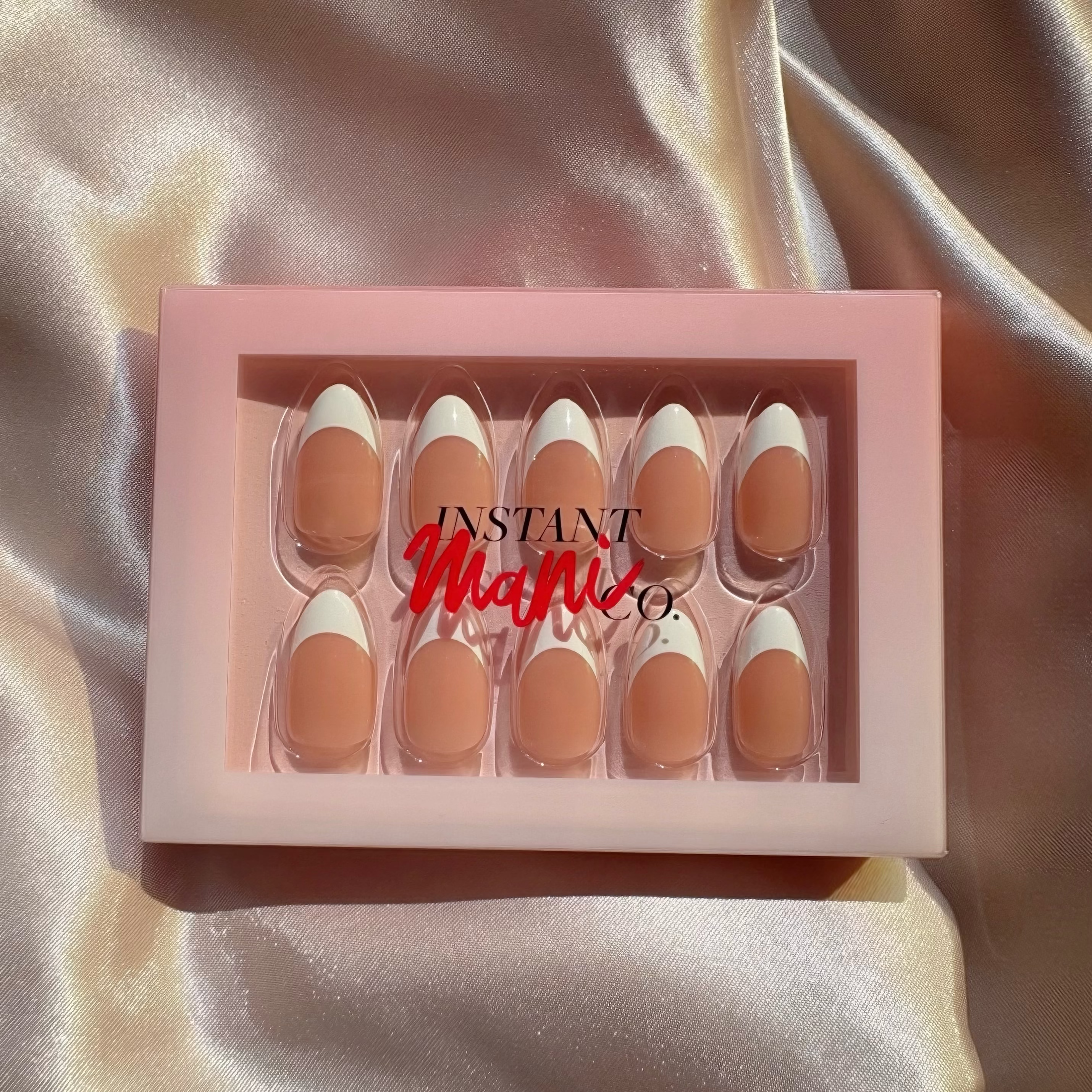 A set of Chic Instant Mani Co. press-on nails in a pink tray with a clear lid, featuring a French tip design with a nude beige base and white tips. The nails are displayed on a shiny, draped champagne-colored satin fabric background.