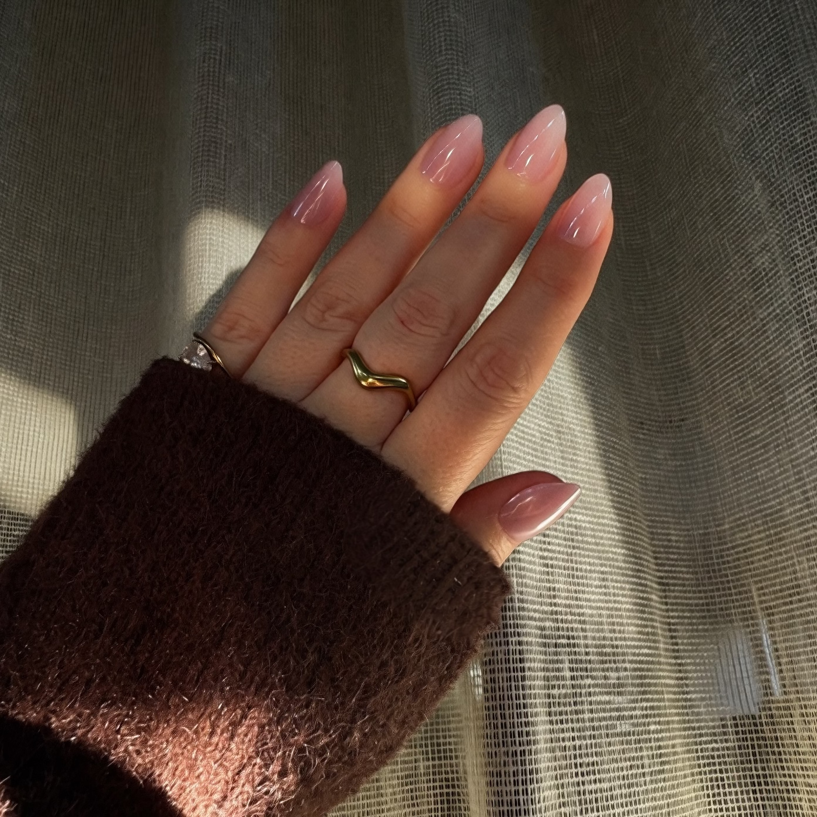 A close-up of a hand with Instant mani Co. almond-shaped press-on nails in a glossy dusty pink shade called Halo, wearing a brown knit sweater sleeve against a background of sheer beige curtains with warm sunlight.
