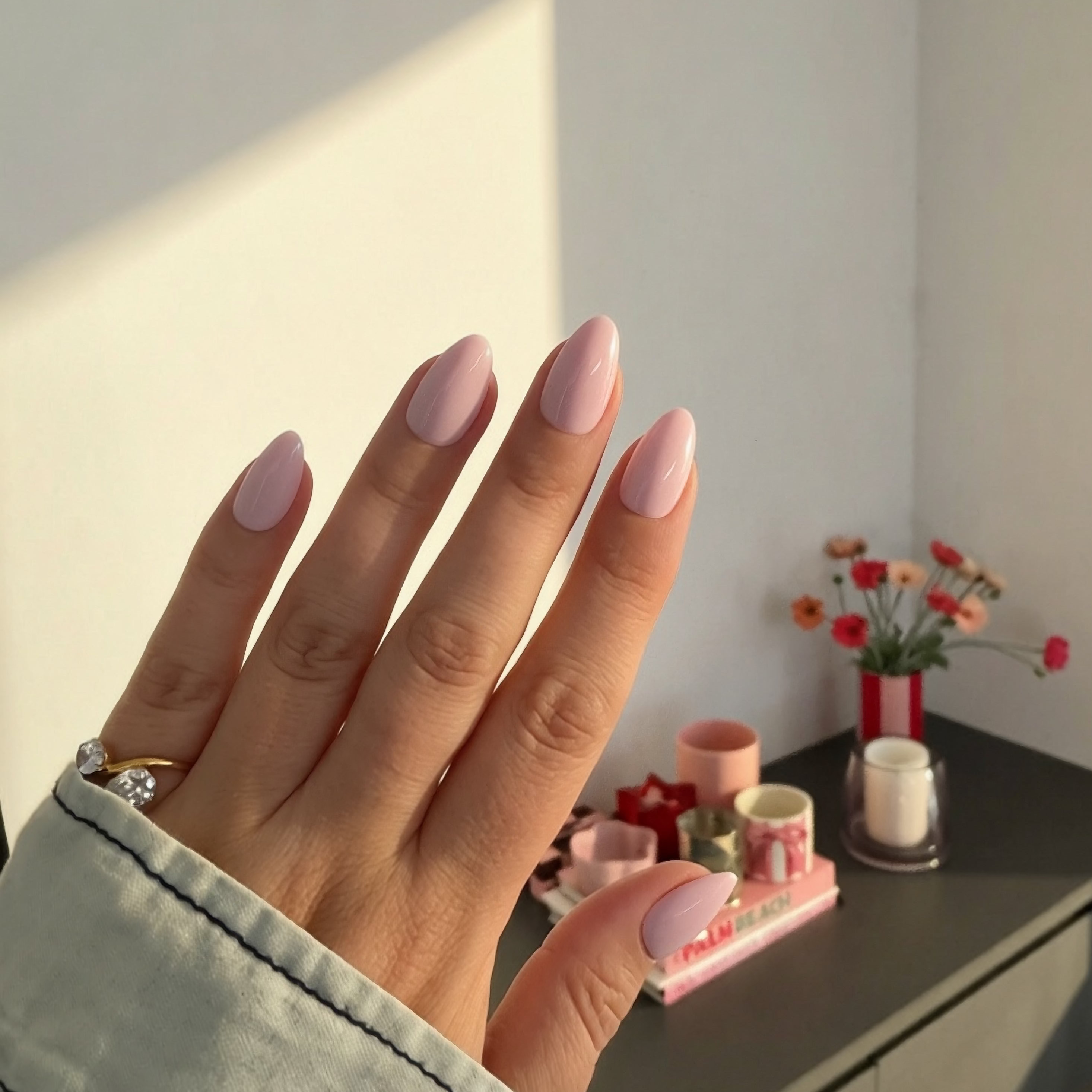 Hand with Instant Mani Co. peony pink  press on nails in a softly lit room with decorative items on a shelf.