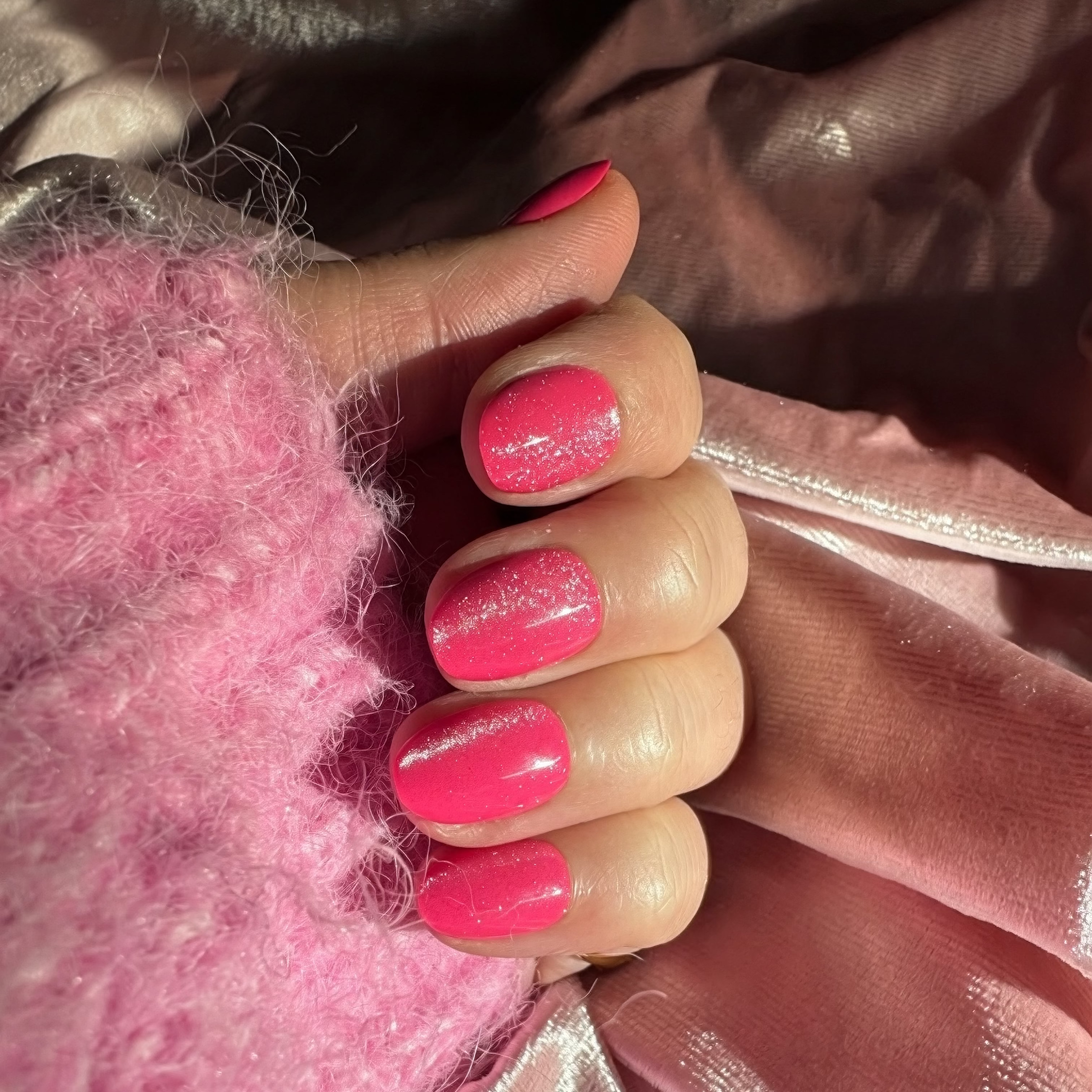 A close-up of a hand with bright velvet Instant Mani Co. press on nails, wearing gold rings and a fuzzy pink sweater, with velvet in the background.