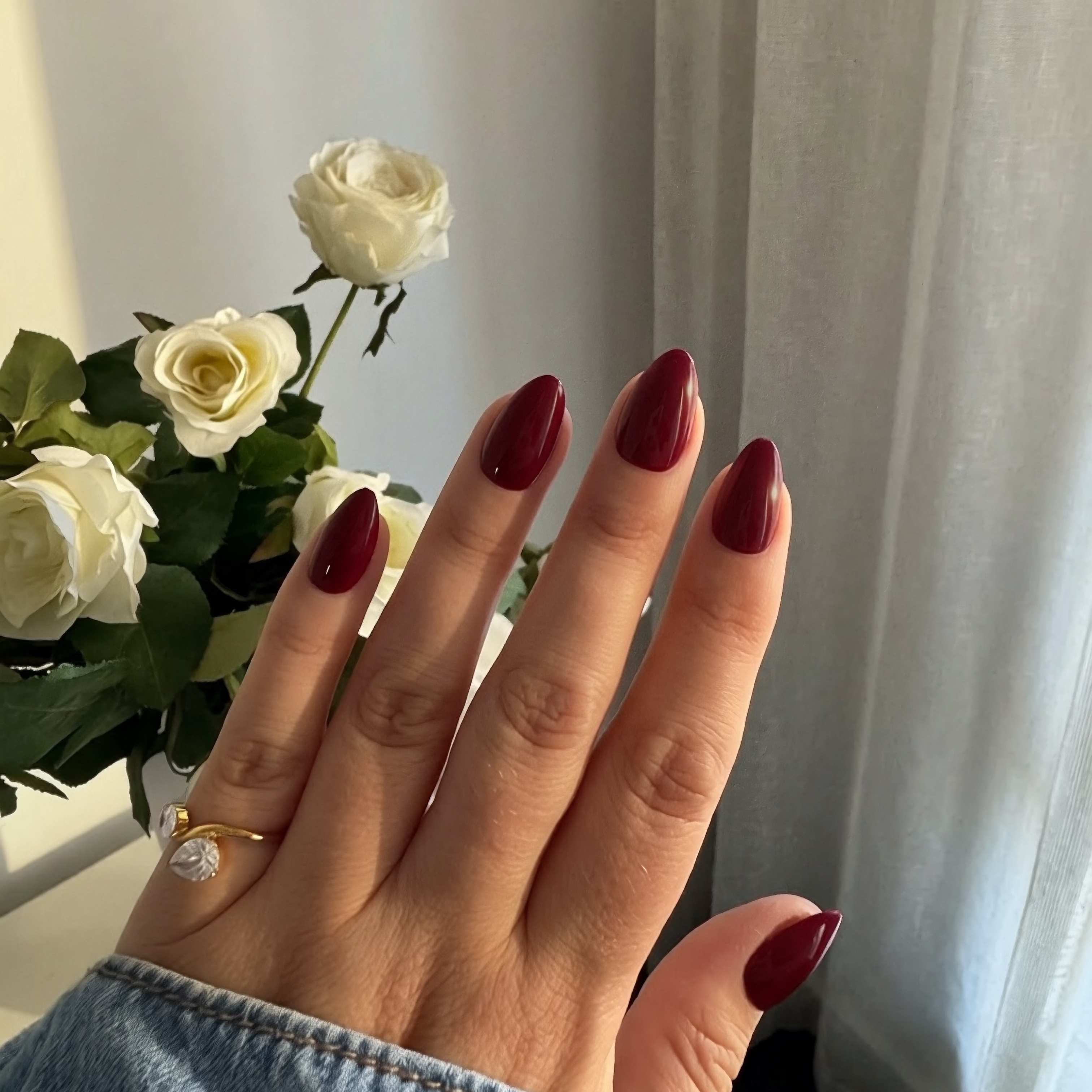 Hand with Instant Mani Co. Rogue Press on Nails wearing a gold ring, holding white roses against a light curtain background