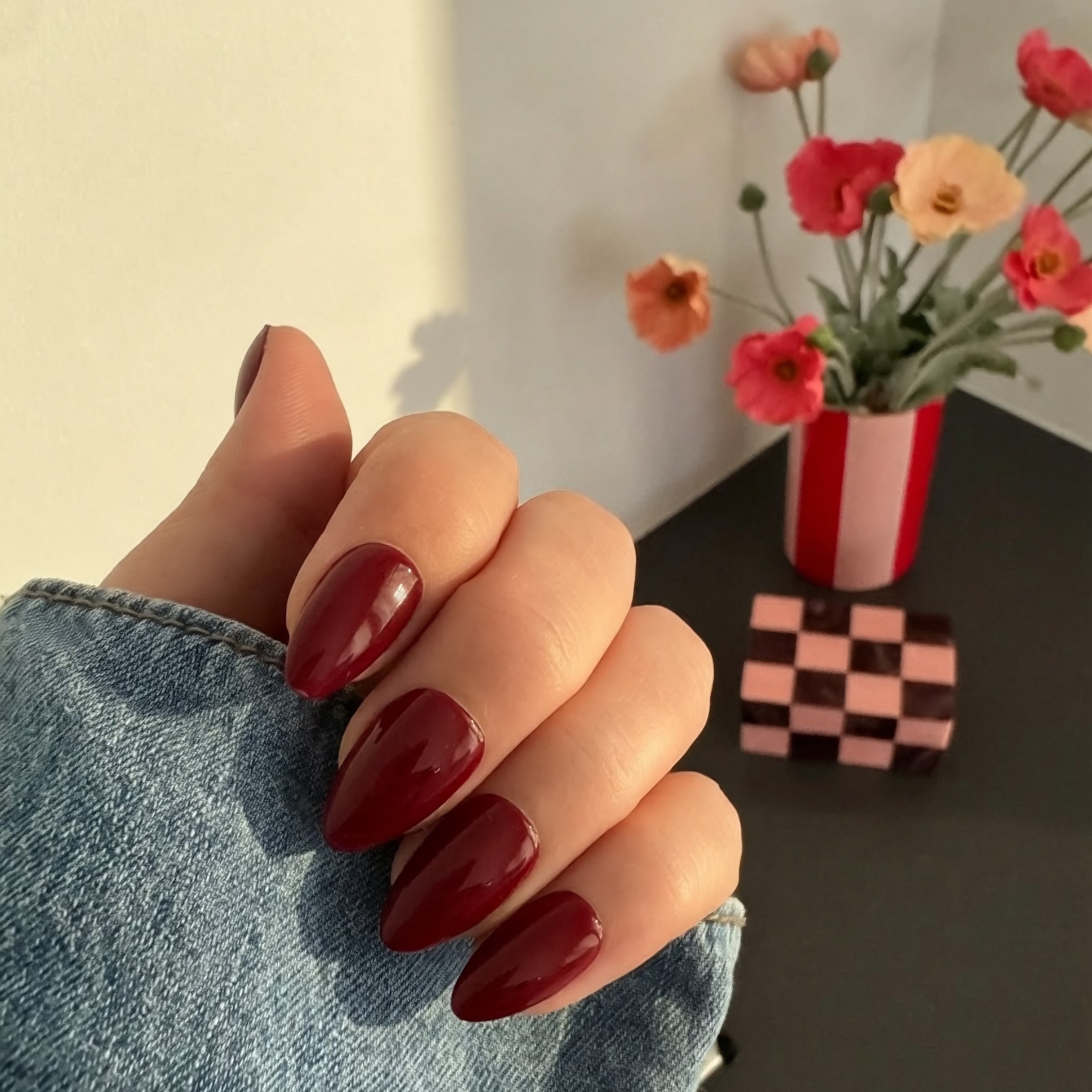 Hand with Instant Mani Co.press on nails wearing a denim jacket, with a vase of flowers in the background.