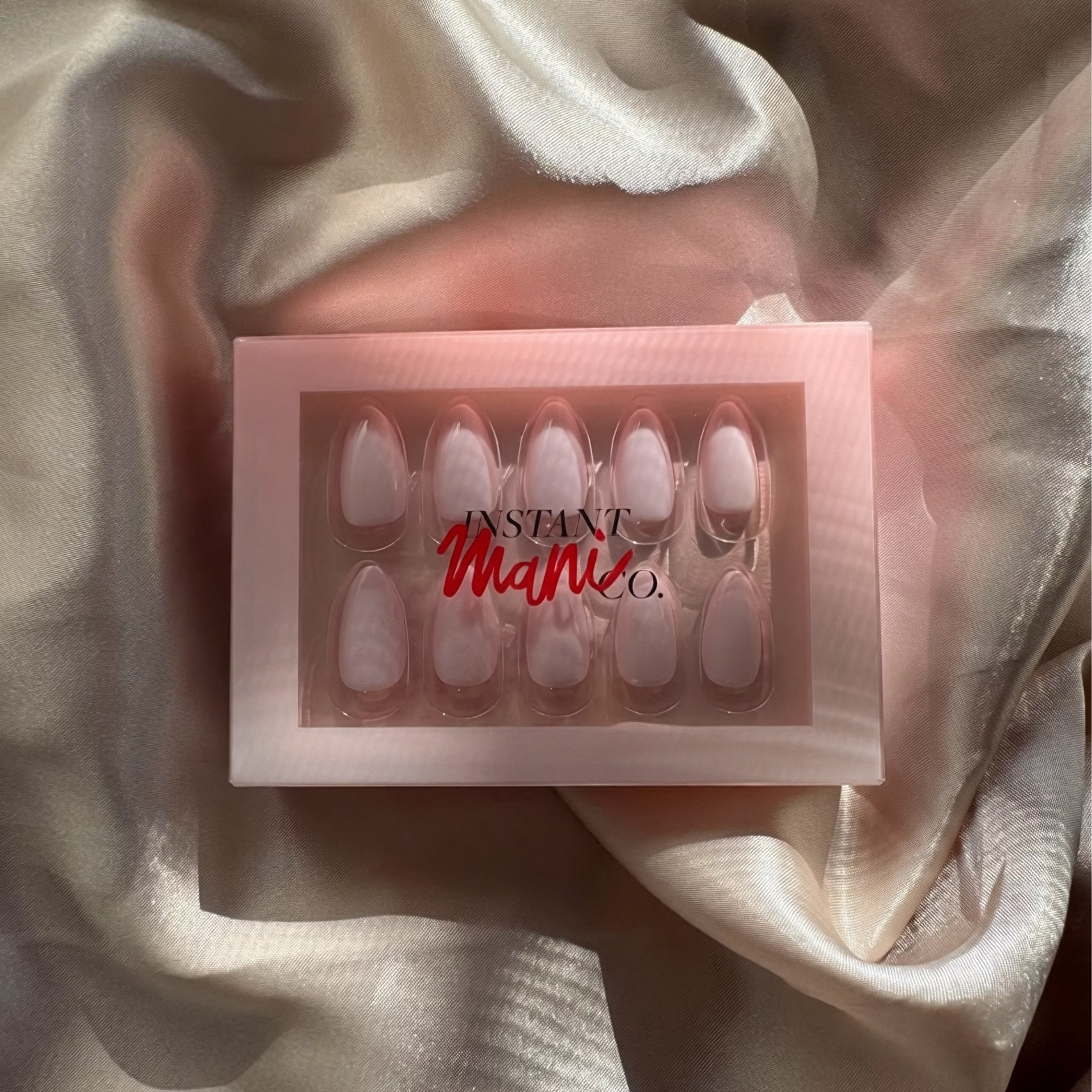 Box of press on nails with 'Instant Mani Co.' branding on a soft fabric background
