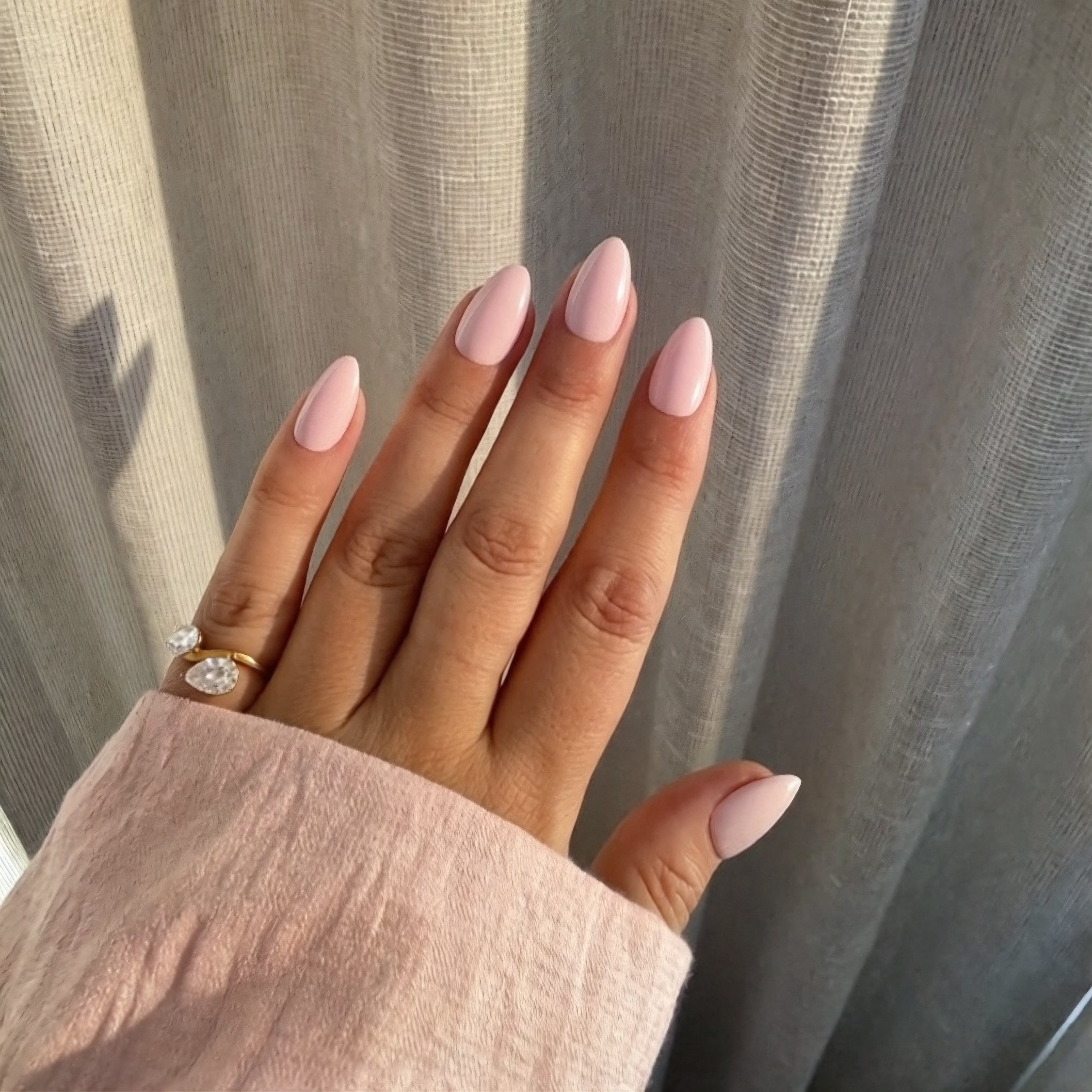 Hand with Instant Mani Co. pink peony press-on nails wearing a ring against a neutral curtain background