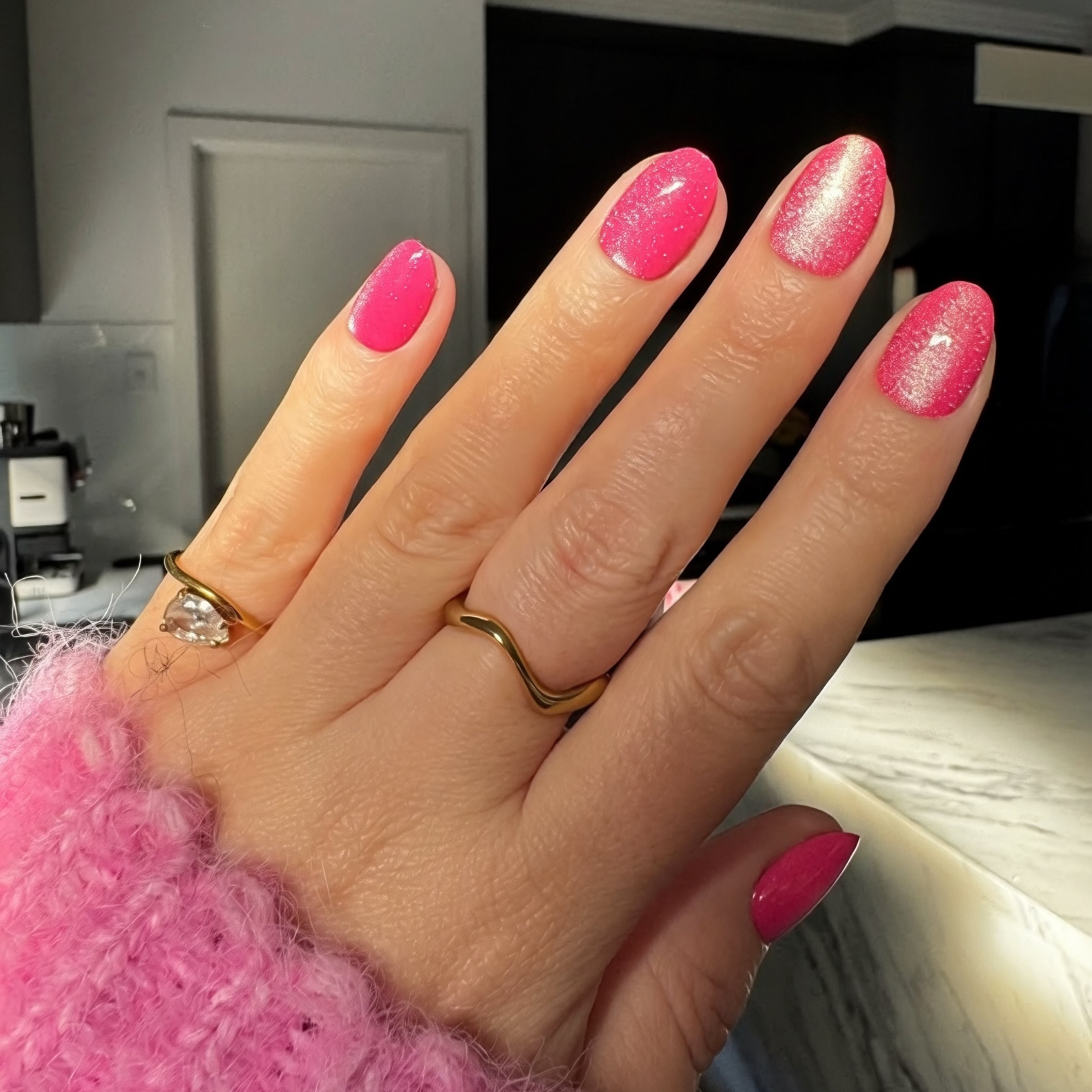 A close-up of a hand with bright velvet Instant Mani Co. press on nails, wearing gold rings and a fuzzy pink sweater, with a kitchen in the background.