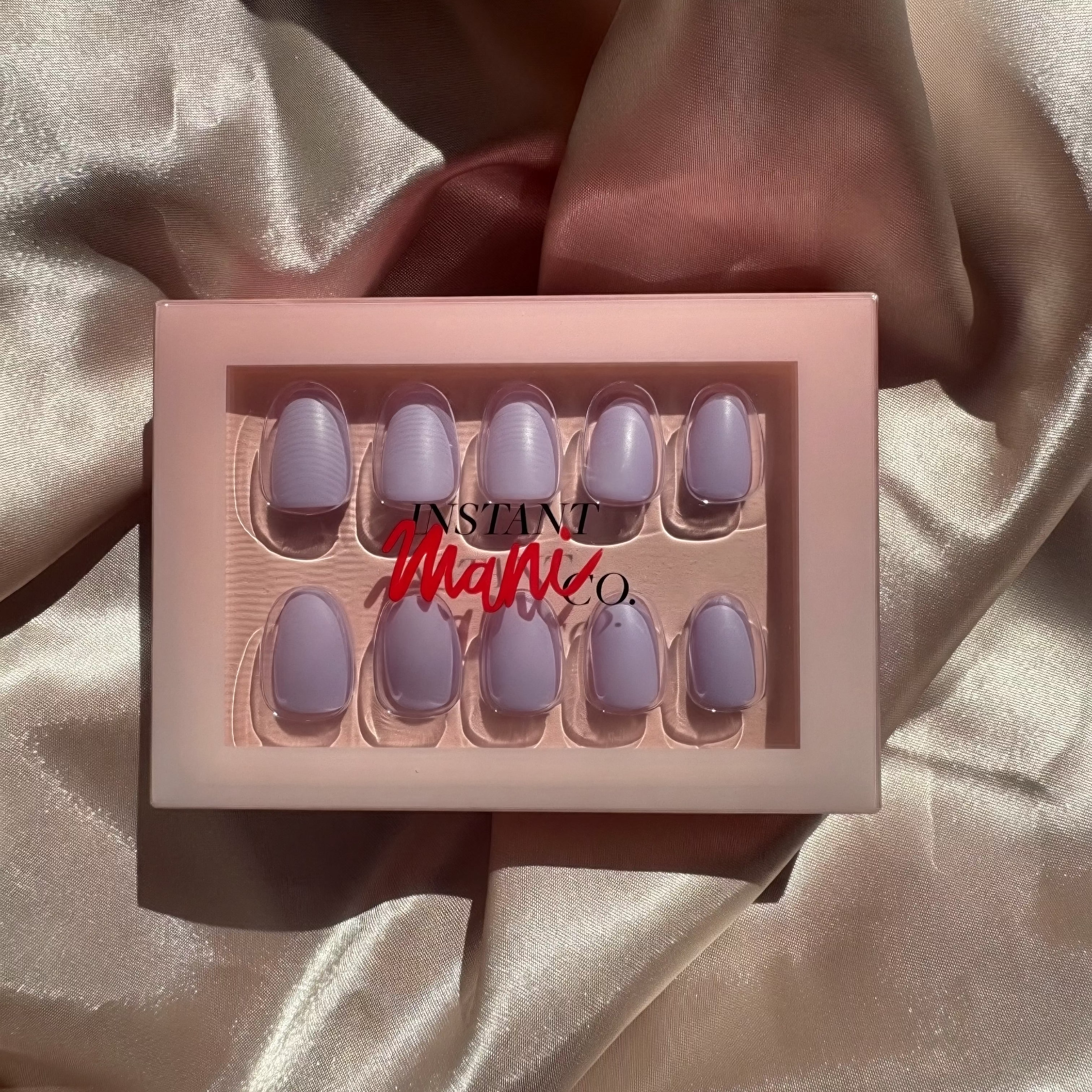 A set of lilac haze press-on nails in a pink tray with the brand name 'INSTANT Mani Co.' printed on the front, resting on draped champagne satin fabric.