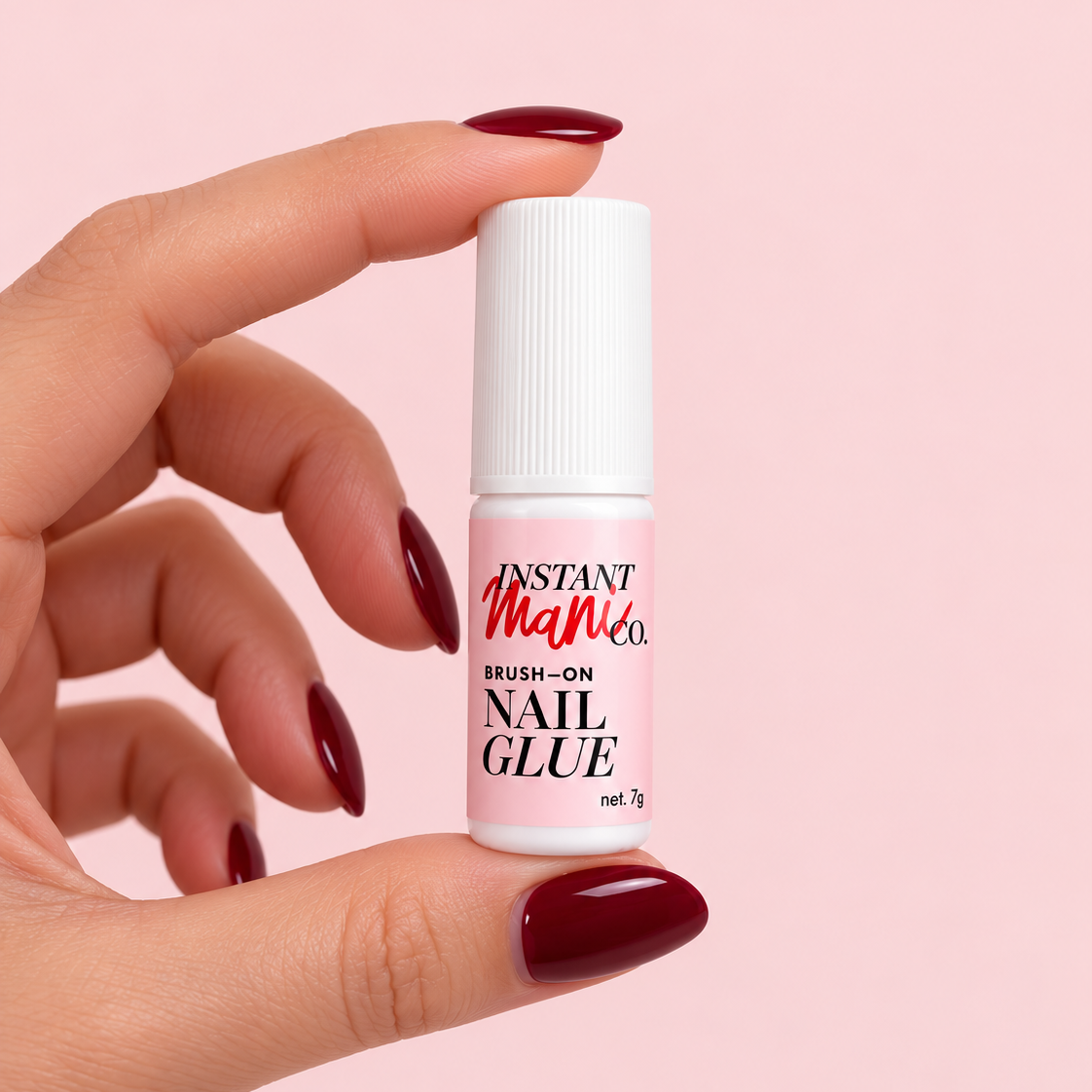 A hand with dark red manicured nails holds a small bottle of Instant Man Co Brush-On Nail Glue against a solid light pink background.