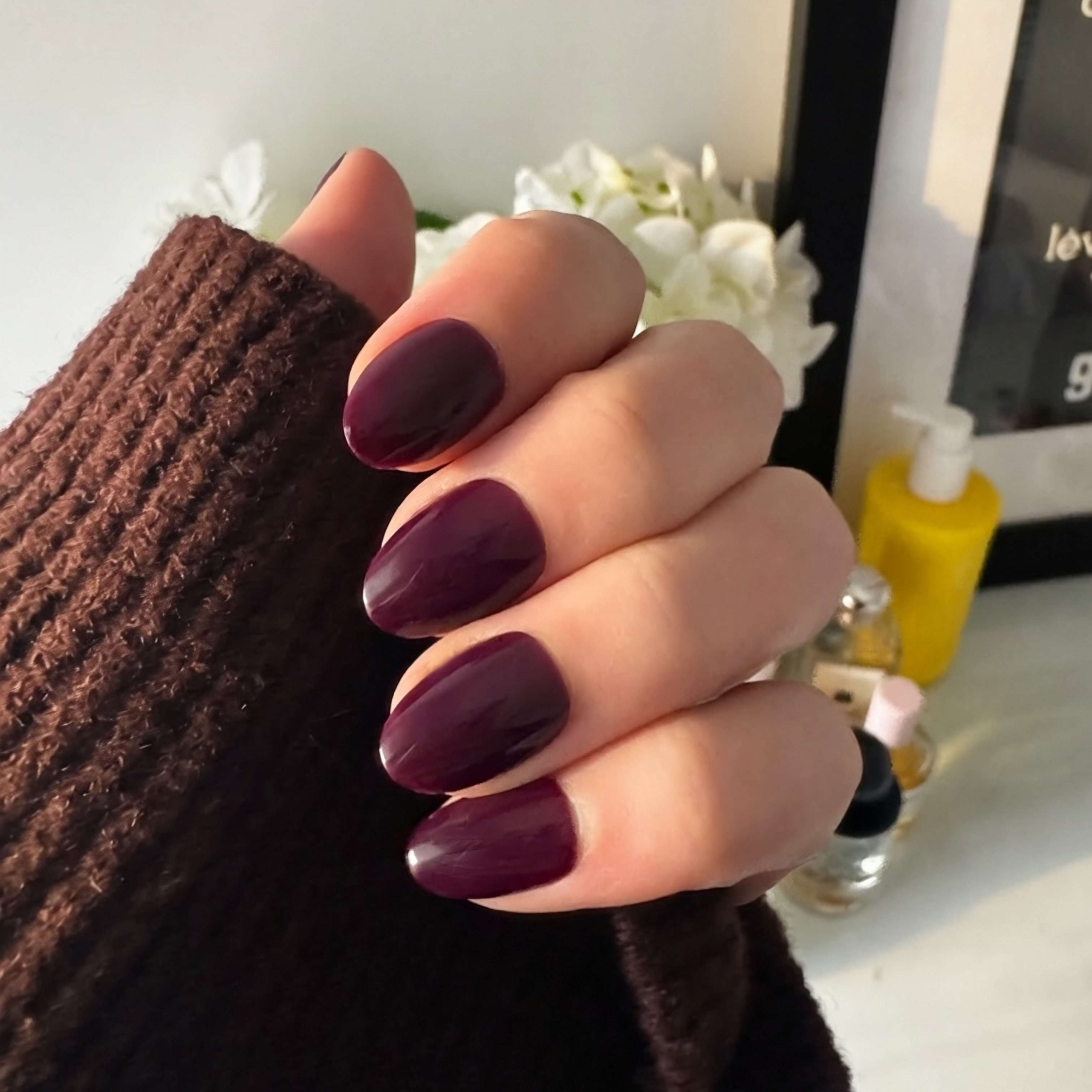 Hand with dark plum Instant Mani Co. press on nails holding a brown textured sleeve, with a blurred background.