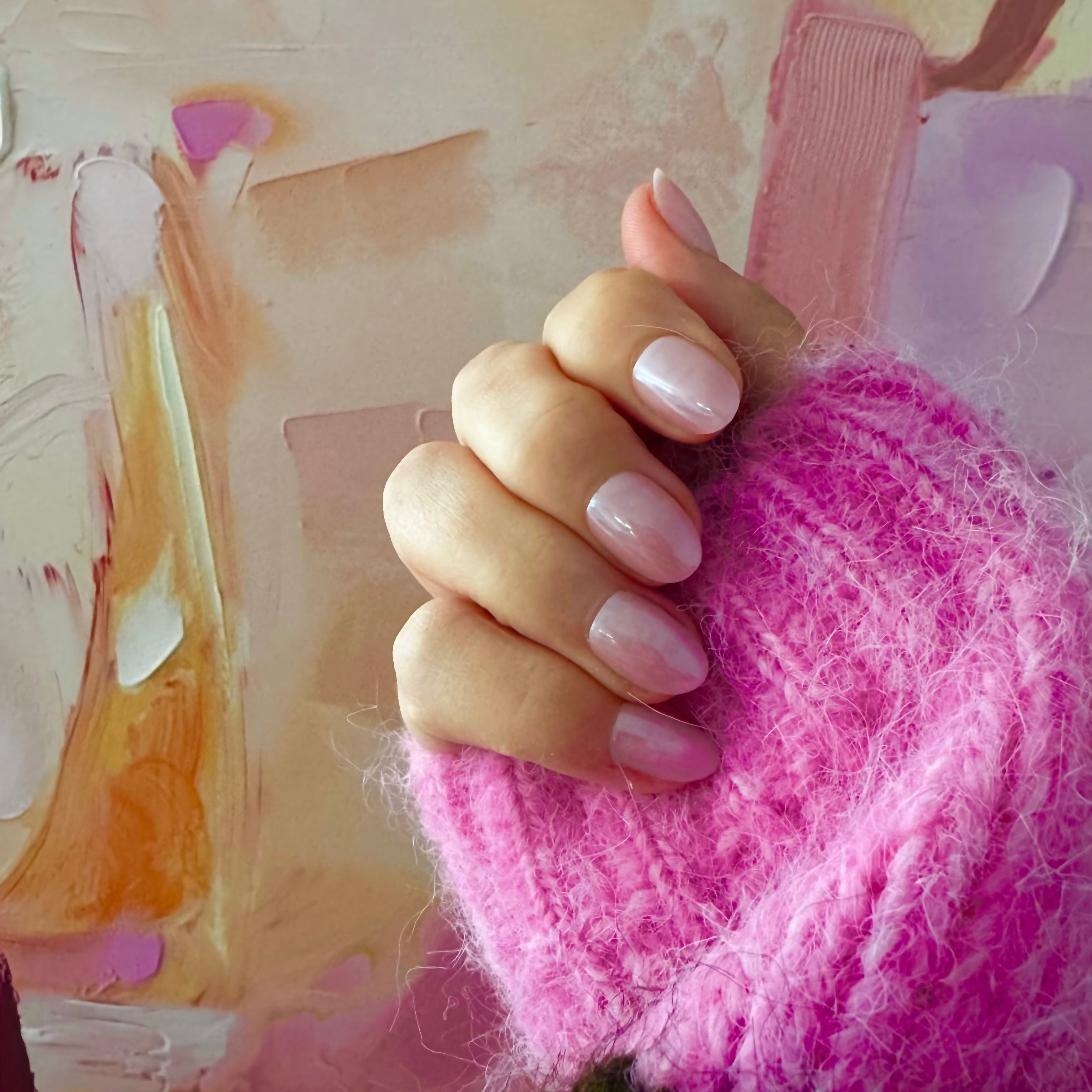 A close-up of a hand wearing glossy pink Instant Mani Co Press on nails in Strawberry Glaze resting on a fluffy bright pink knit sleeve, set against an abstract pastel painted background.