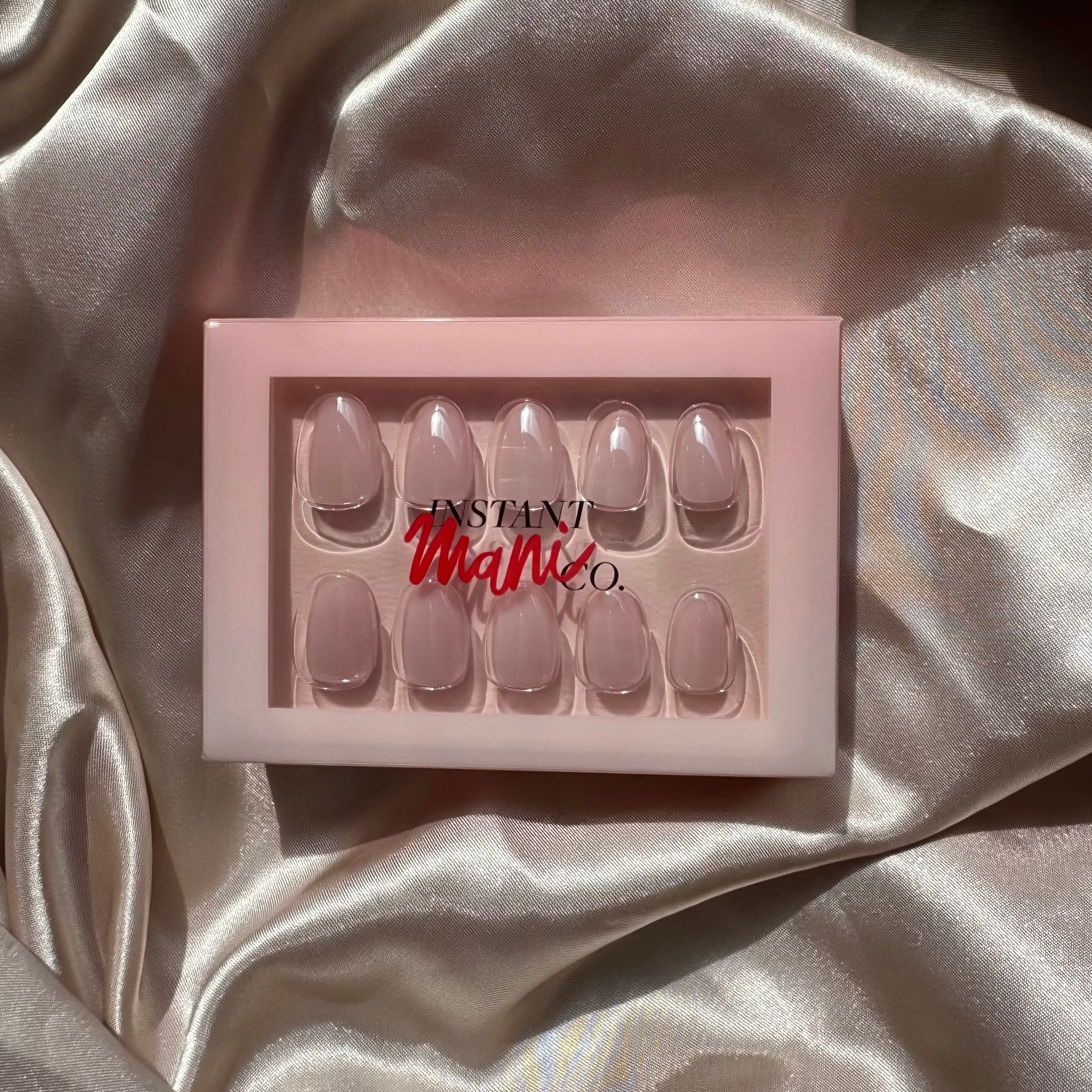A set of 10 press-on nails in a pale pink shade, presented in a light pink box with the brand name 'Instant Mani co' visible, set against a draped champagne satin fabric background.