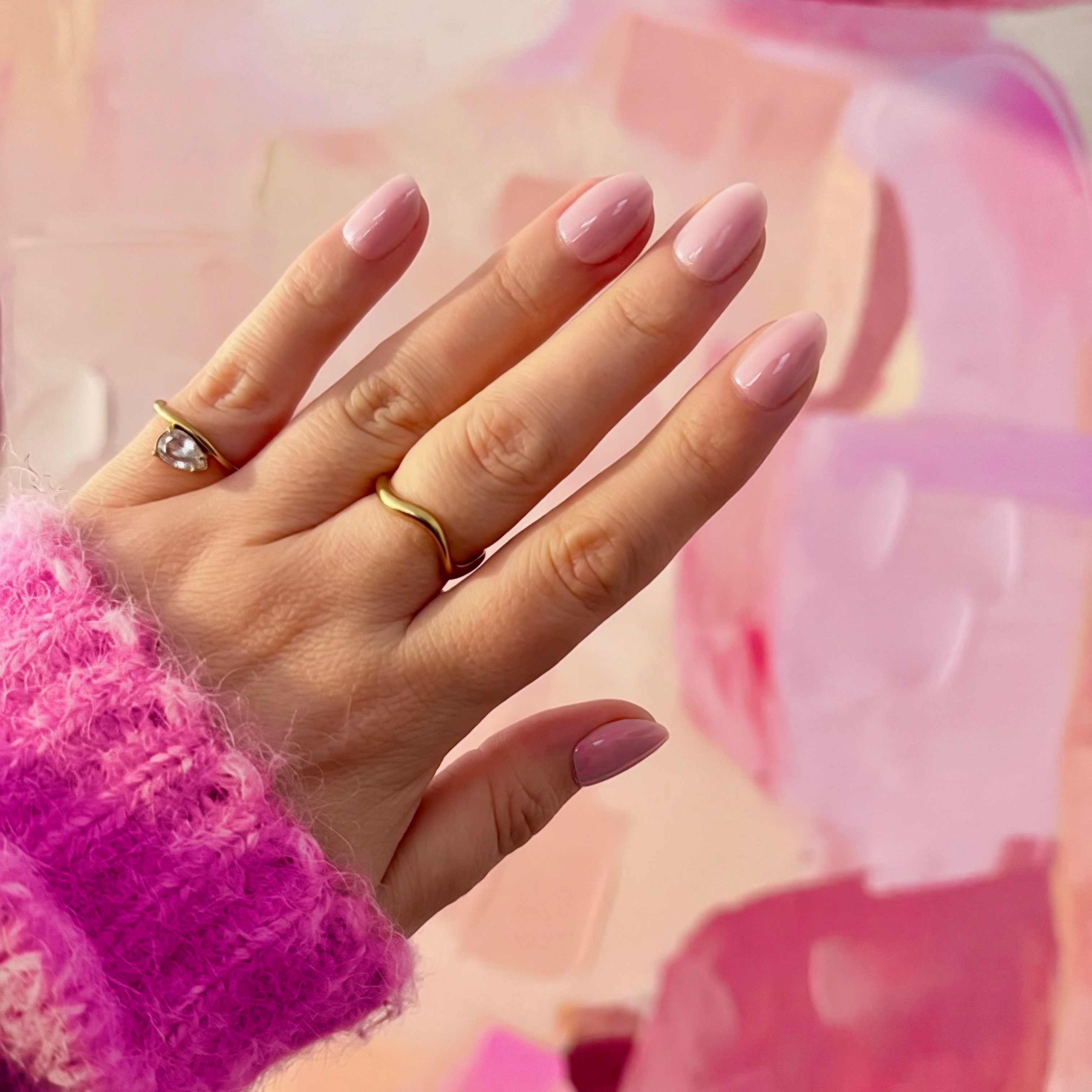 A close-up of a hand with light pink Instant Mani Co. Press on Nails, wearing a gold band ring and a silver ring with a clear stone, set against a soft pink and peach abstract painted background.