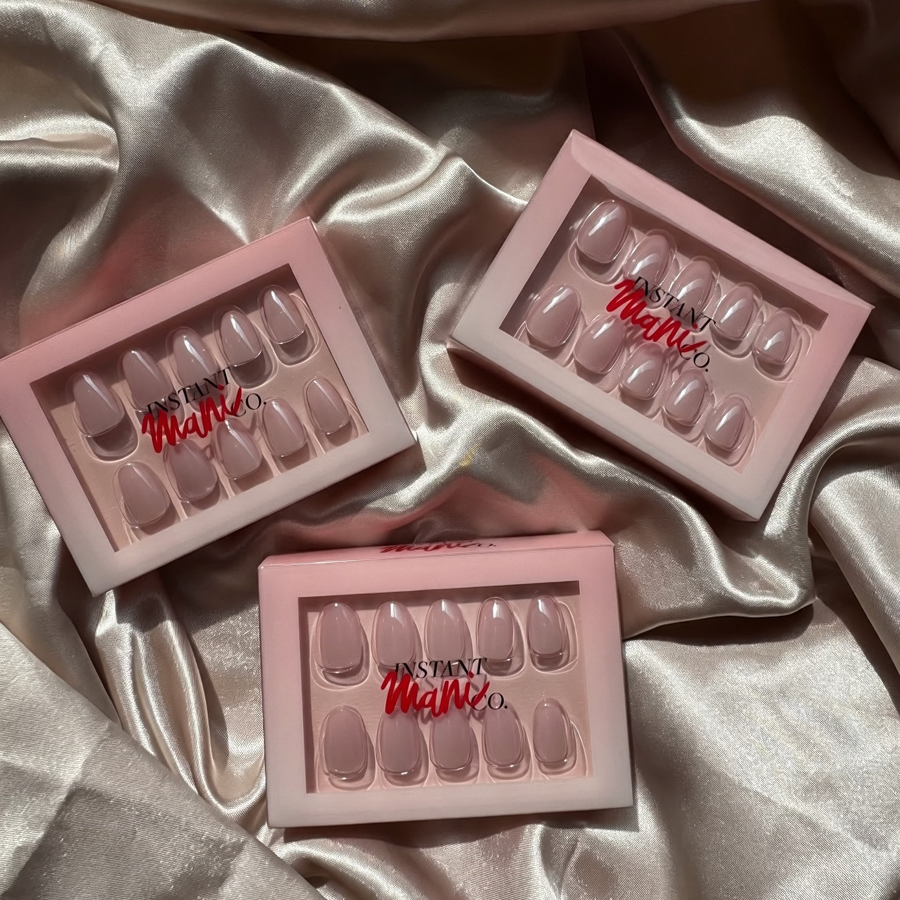Three pink Instant Magi co press-on nail kits arranged on a draped champagne satin fabric background.