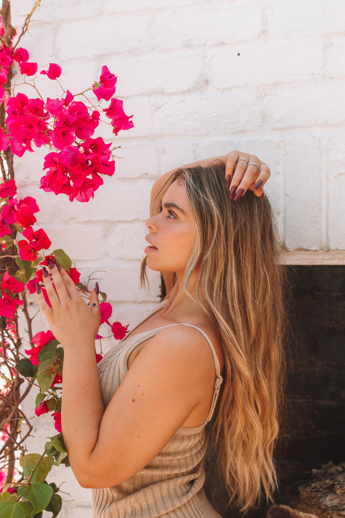 girl wearing Instant Mani press-on nails in burgundy, looking at pink plant. 