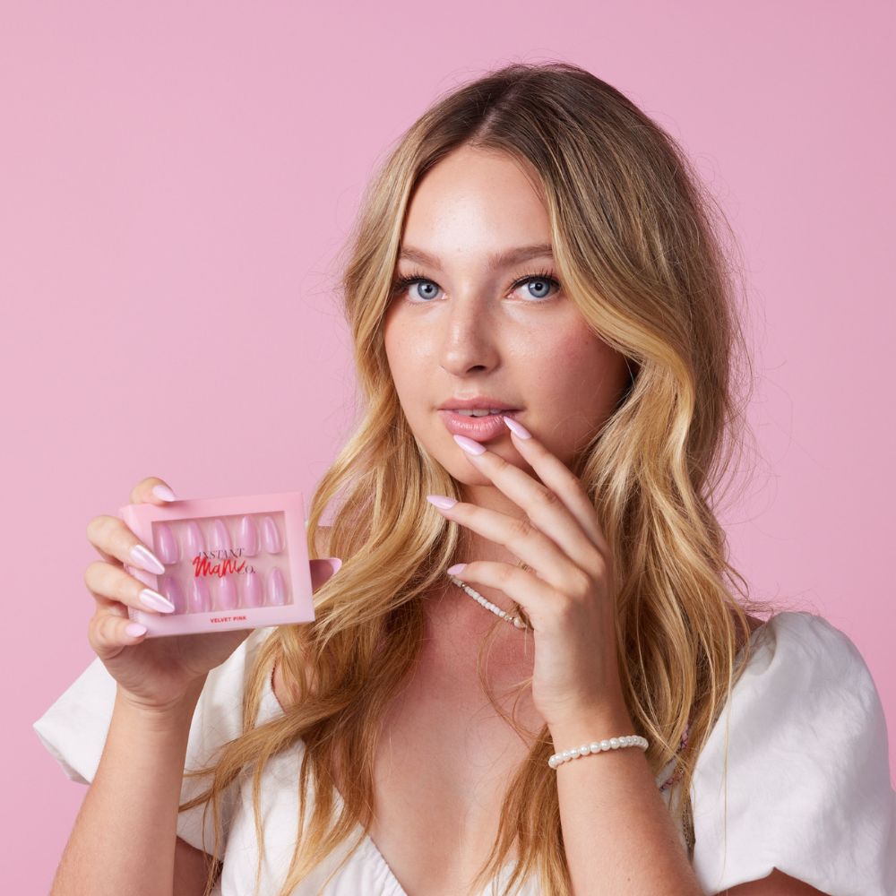 Girl wearing Instant Mani Co Velvet Pink Press on nails, holding Instant Mani Co. box,  standing in front of pink background wearing white shirt 