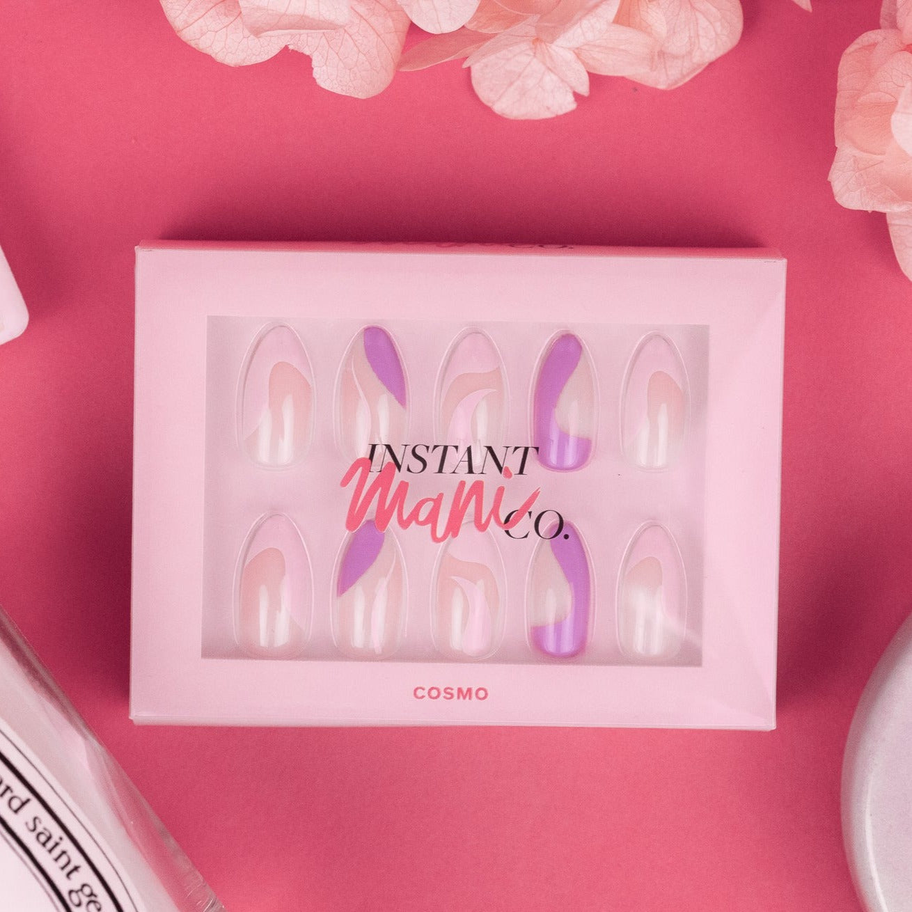 Box of Instant Mani Co. Cosmo press-on nails on pink background with brush-on nail glue and flowers in shot.