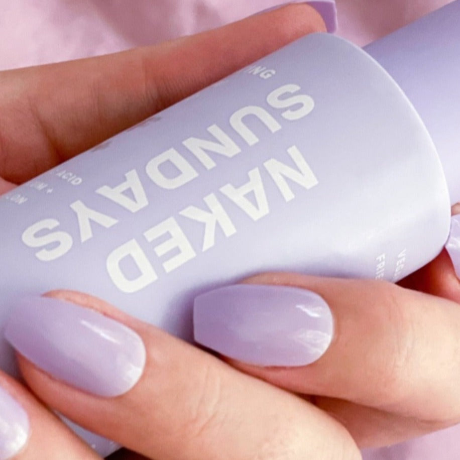 hand wearing Instant Mani Pastel Purple press-on nails holding bottle of purple Naked Sundays sunscreen