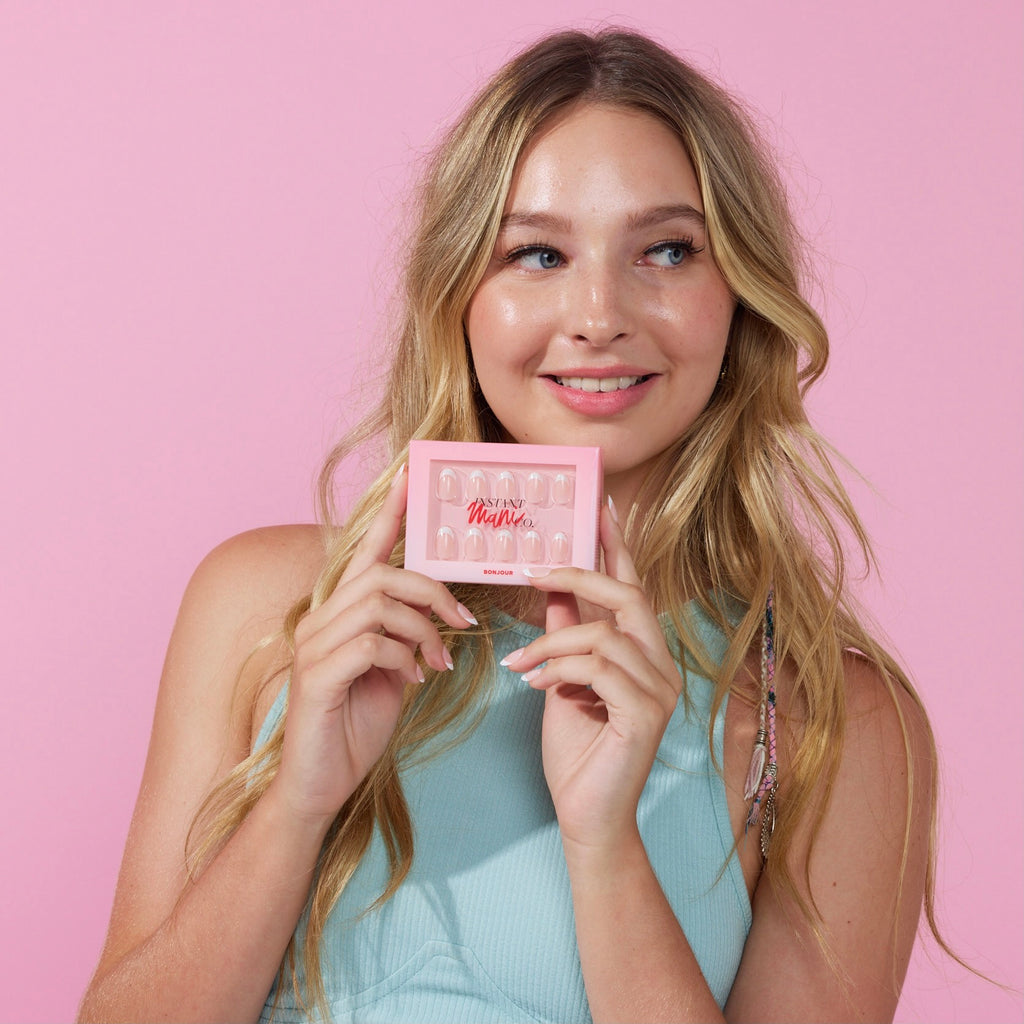 Girl wearing Instant Mani Bonjour press on nails standing in front of pink background wearing blue shirt