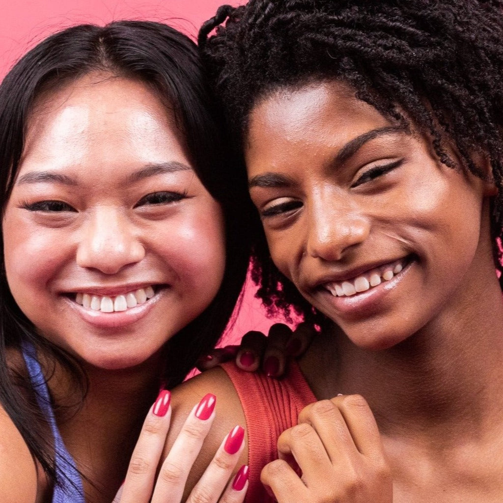 Two girls smiling, in front of a pink background, one wearing red Instant Mani Co. press-on nails