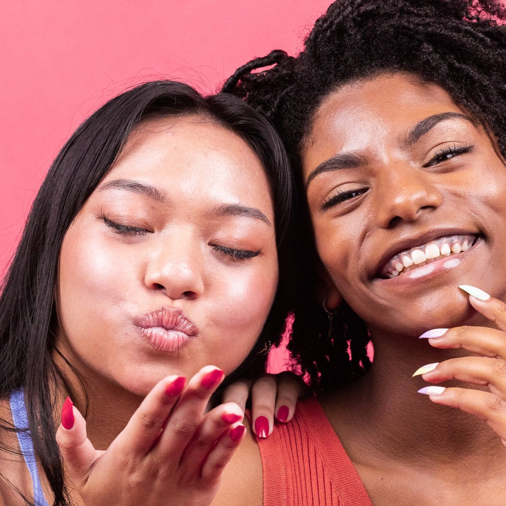 two girls smiling, one blowing a kiss, standing in front of a pink background, wearing Instant Mani Co. press-on nails
