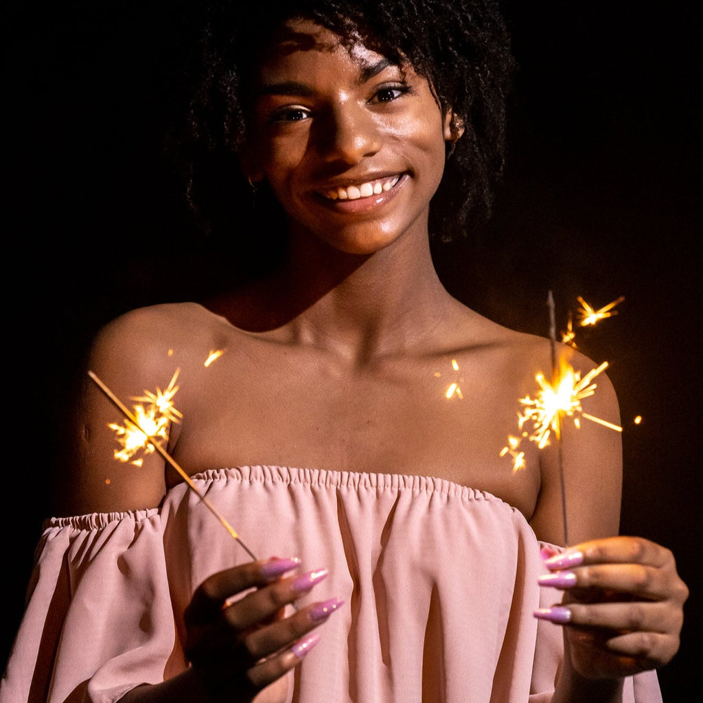Girl wearing pink jumpsuit, standing in front of dark background, holding two sparklers, wearing Instant Mani Co. Stardust, pink glitter press-on nails